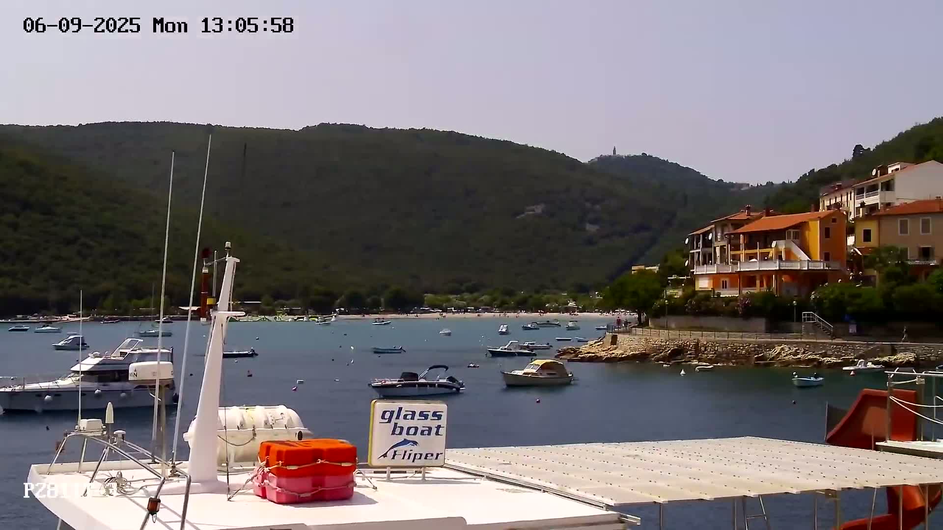 A sunny day shows a calm bay with numerous boats, a small beach with people, and colorful buildings on a hillside overlooking the water.