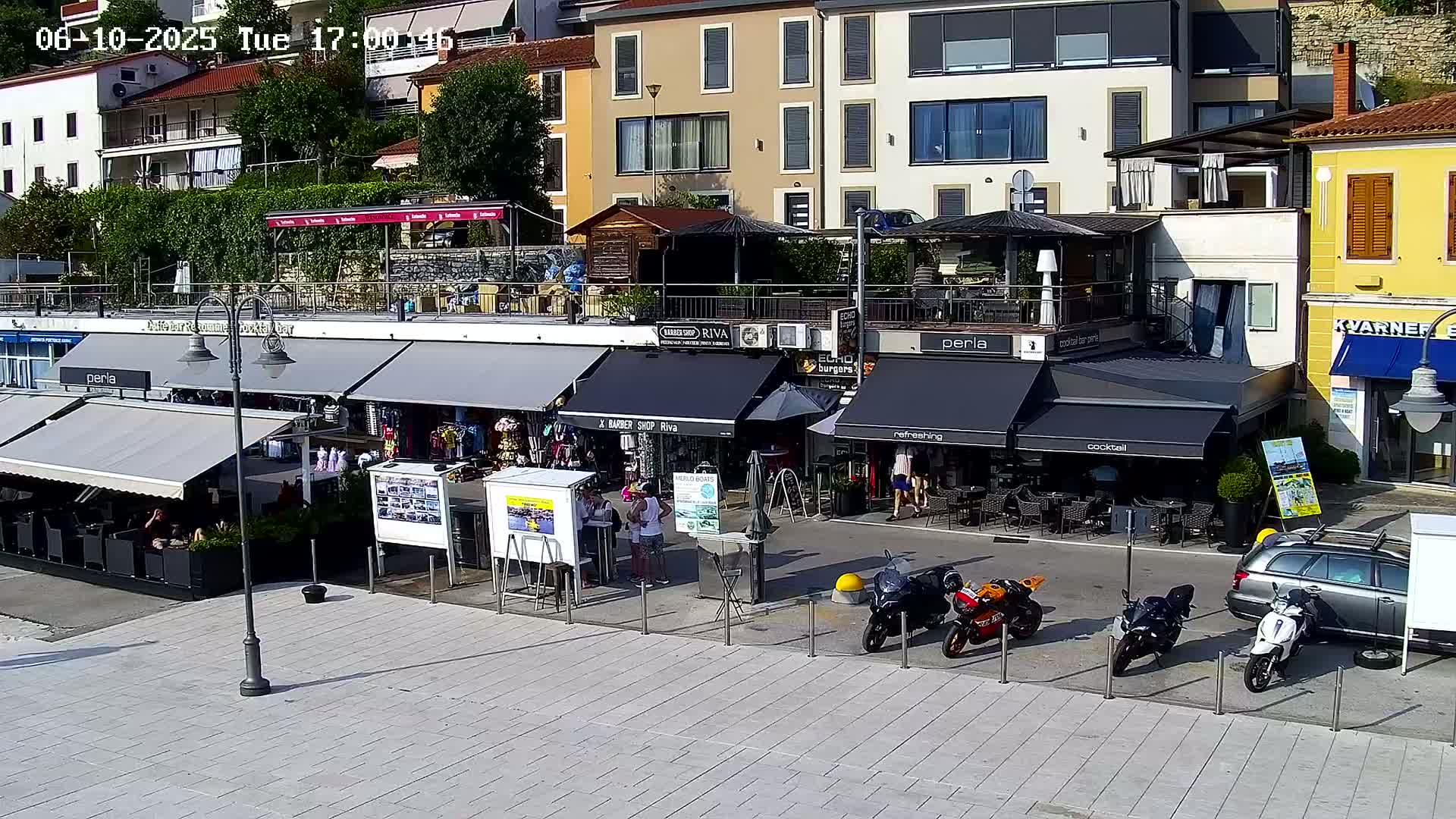 A sunny outdoor scene shows a waterfront area with several shops and restaurants, outdoor seating, and motorcycles parked nearby.