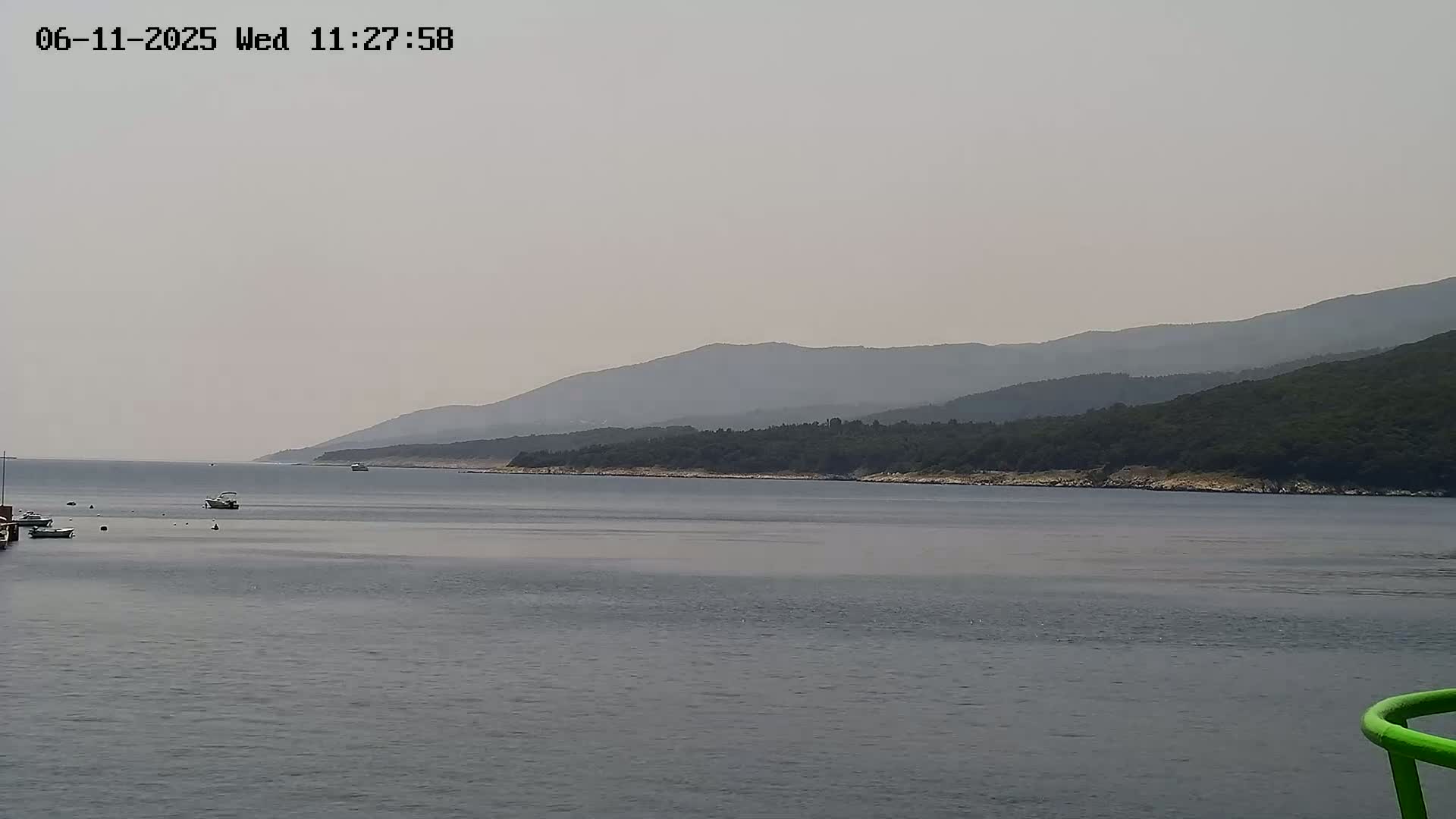 A hazy, overcast day reveals a calm body of water with several small boats, overlooking a shoreline lined with dark green trees and mountains in the distance.