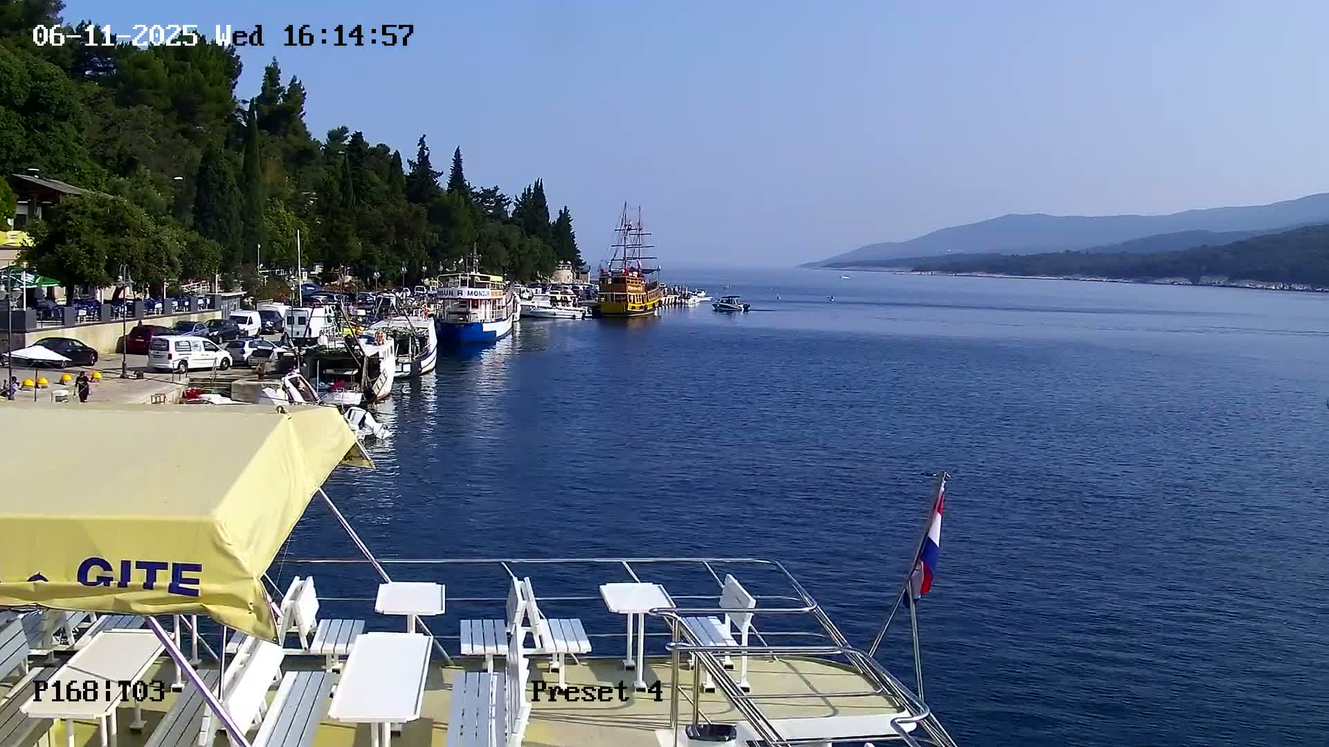 A calm, sunny day shows a harbor filled with boats, with a larger vessel in the foreground,  and mountains in the distance.