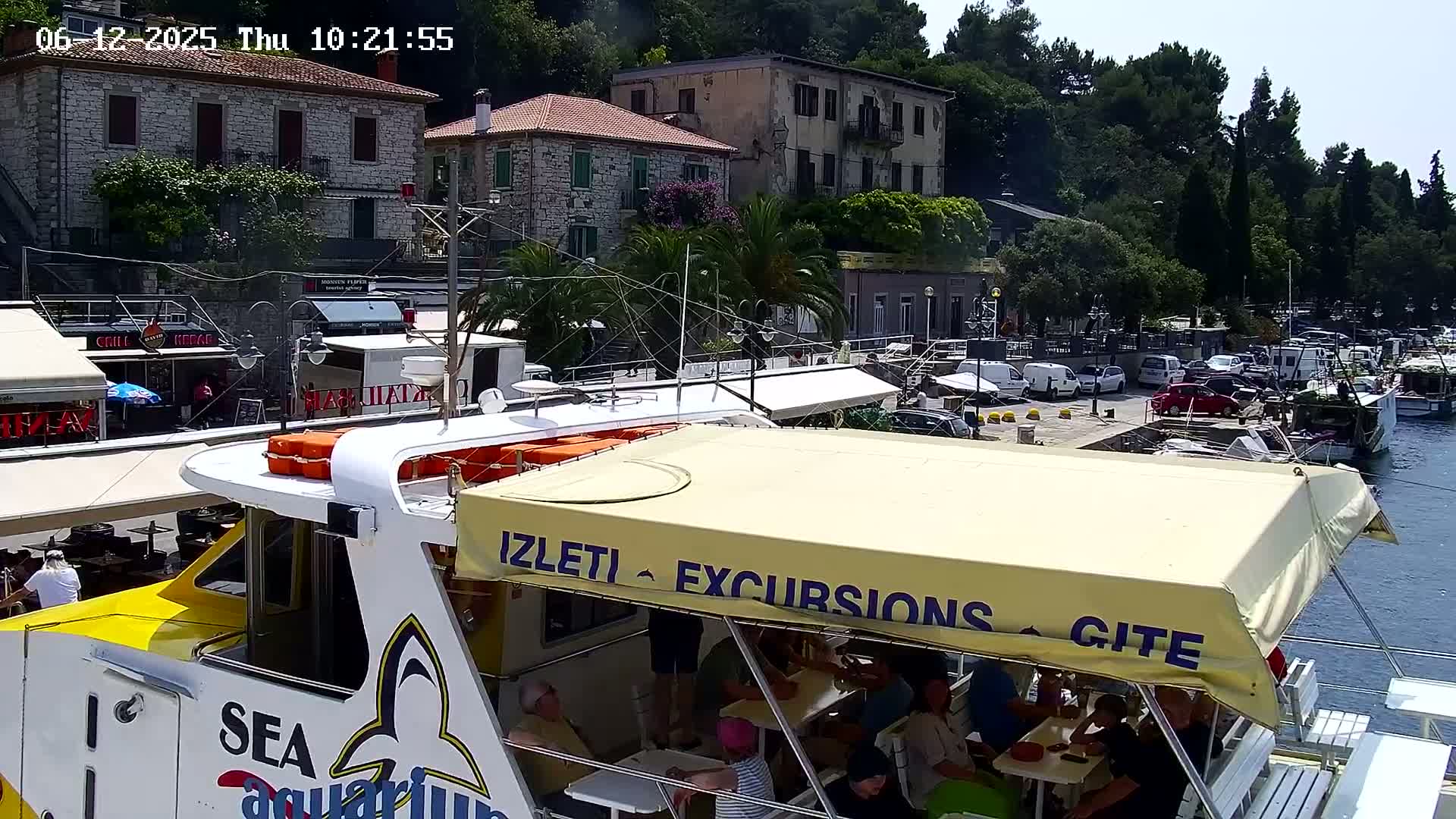 A yellow and white excursion boat with passengers seated under a canopy is docked at a harbor next to stone buildings and parked cars on a sunny day.