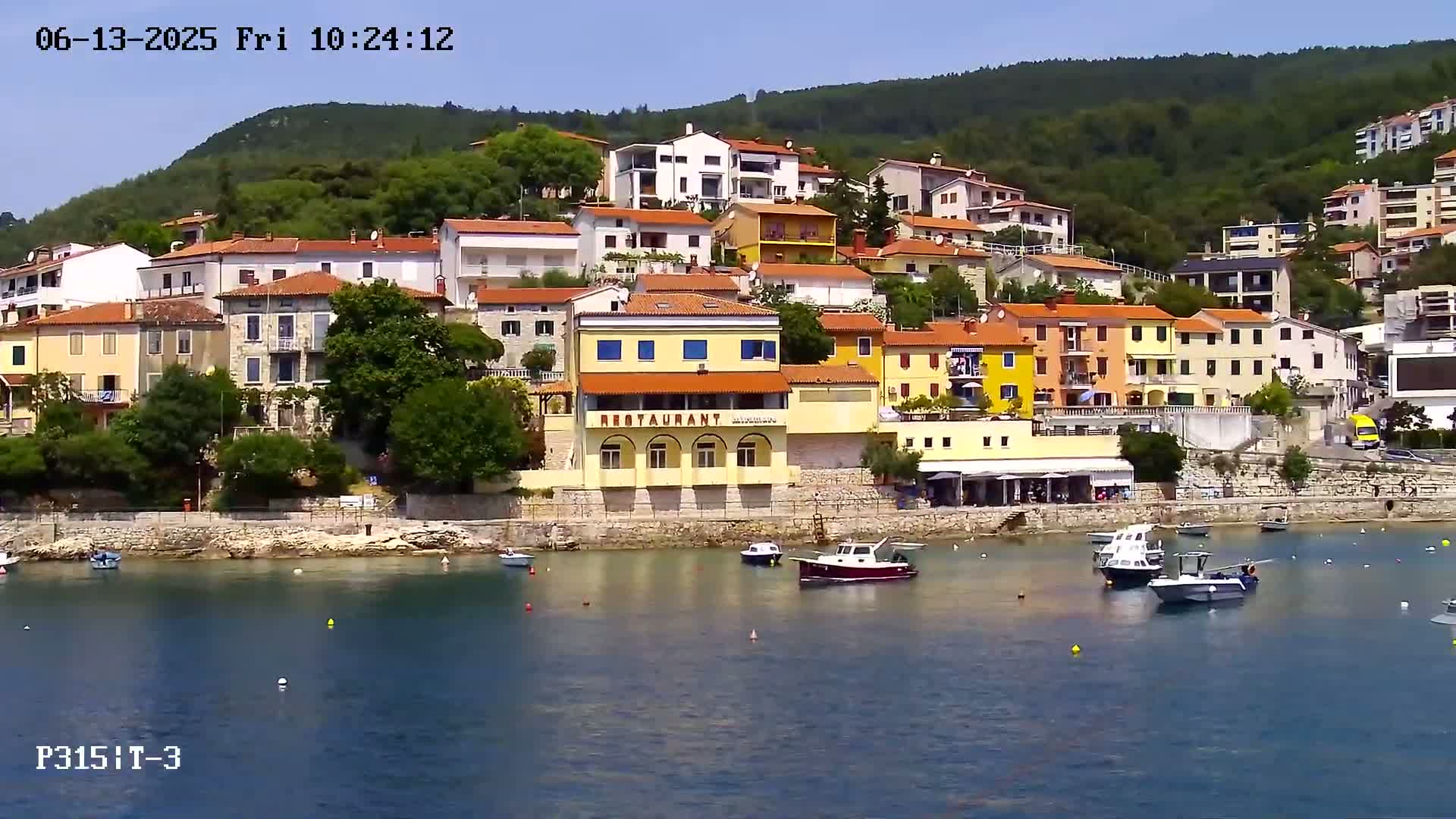 A waterfront village of colorful buildings nestled against a green hillside on a sunny day, with several boats moored in the calm bay.