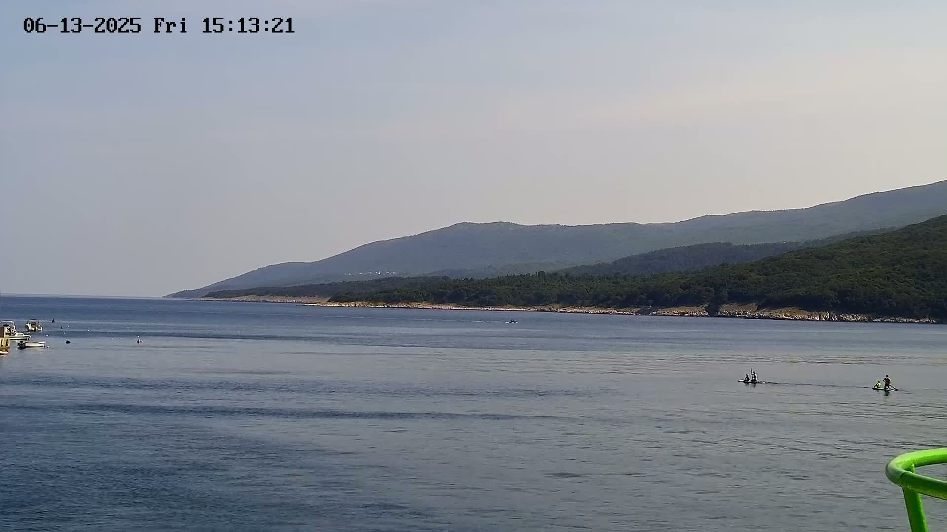 A calm sea with several people paddleboarding near a coastline backed by a mountain range under a clear sky.