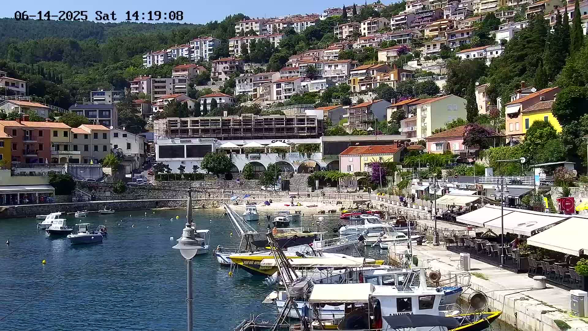 A small harbor filled with boats is nestled beside a hillside town of multi-story buildings under a clear, sunny sky.