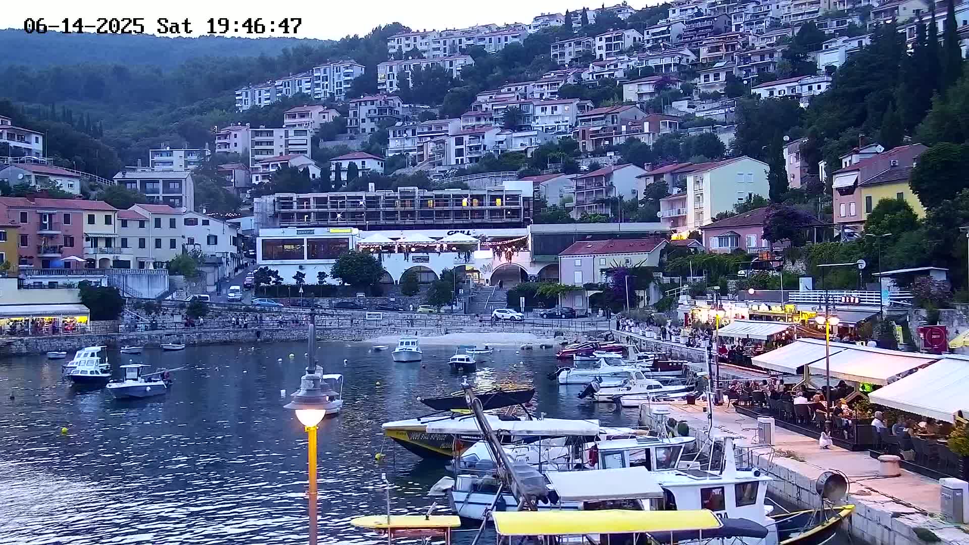 A small harbor filled with boats is nestled beside a hillside town of multi-story buildings under a clear, sunny sky.