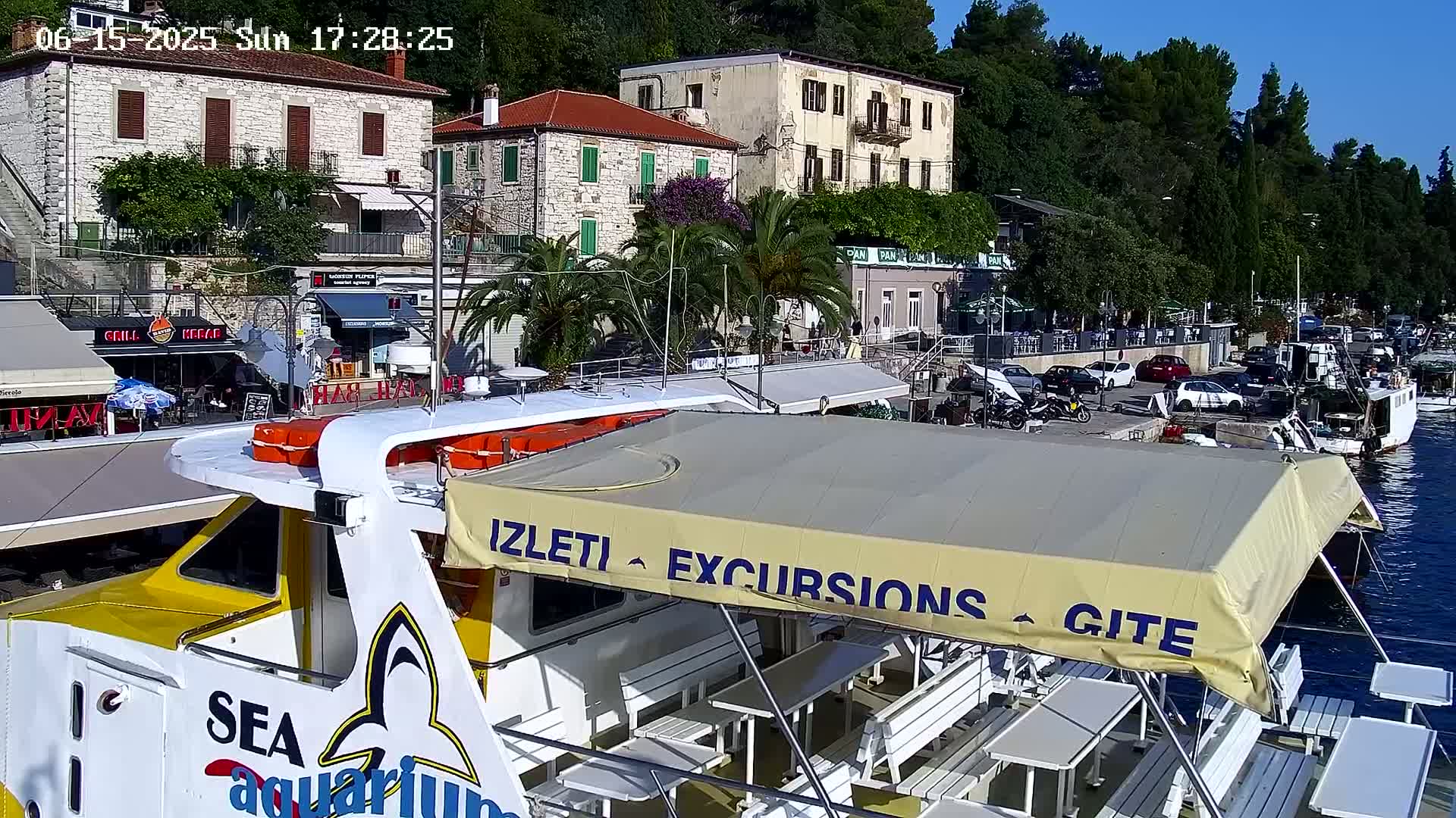 A yellow and white excursion boat is docked at a harbor on a sunny day, next to stone buildings and parked cars.