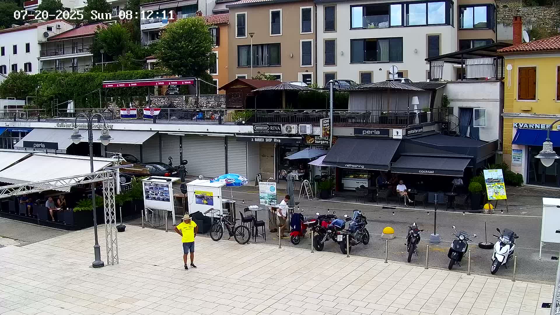 A paved plaza with several outdoor cafes and shops, motorcycles parked along the street, and a person standing in a yellow shirt; the weather appears to be partly sunny.
