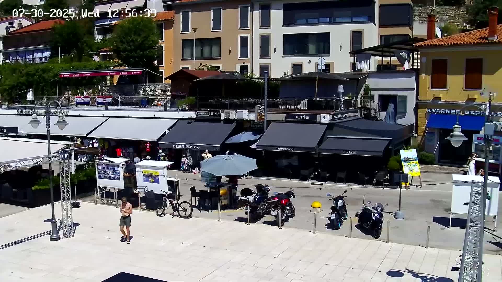 A paved plaza with several outdoor cafes and shops, motorcycles parked along the street, and a person standing in a yellow shirt; the weather appears to be partly sunny.
