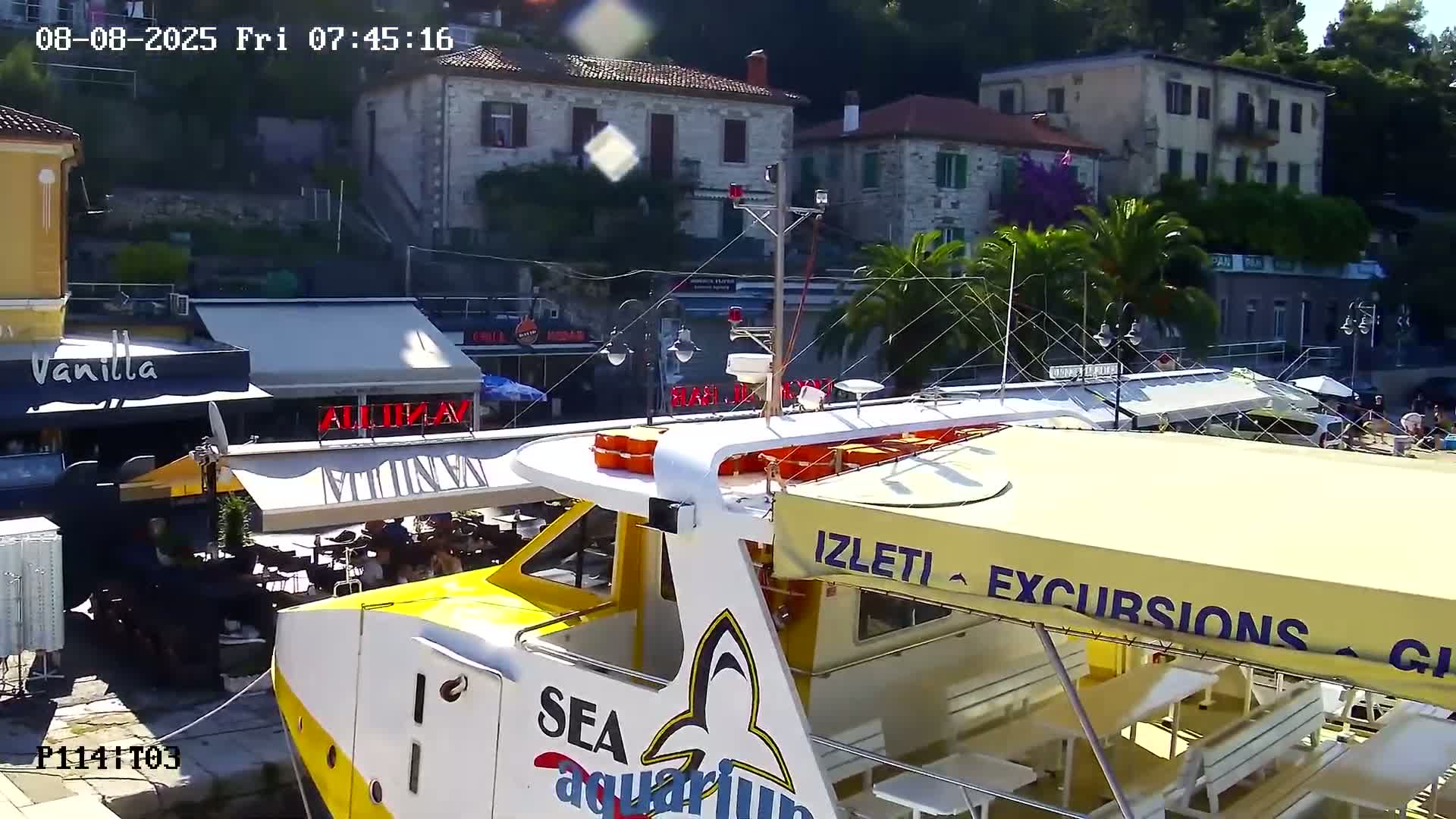 A yellow and white excursion boat is docked at a waterfront, with stone buildings and outdoor cafes visible in the background on a sunny day.