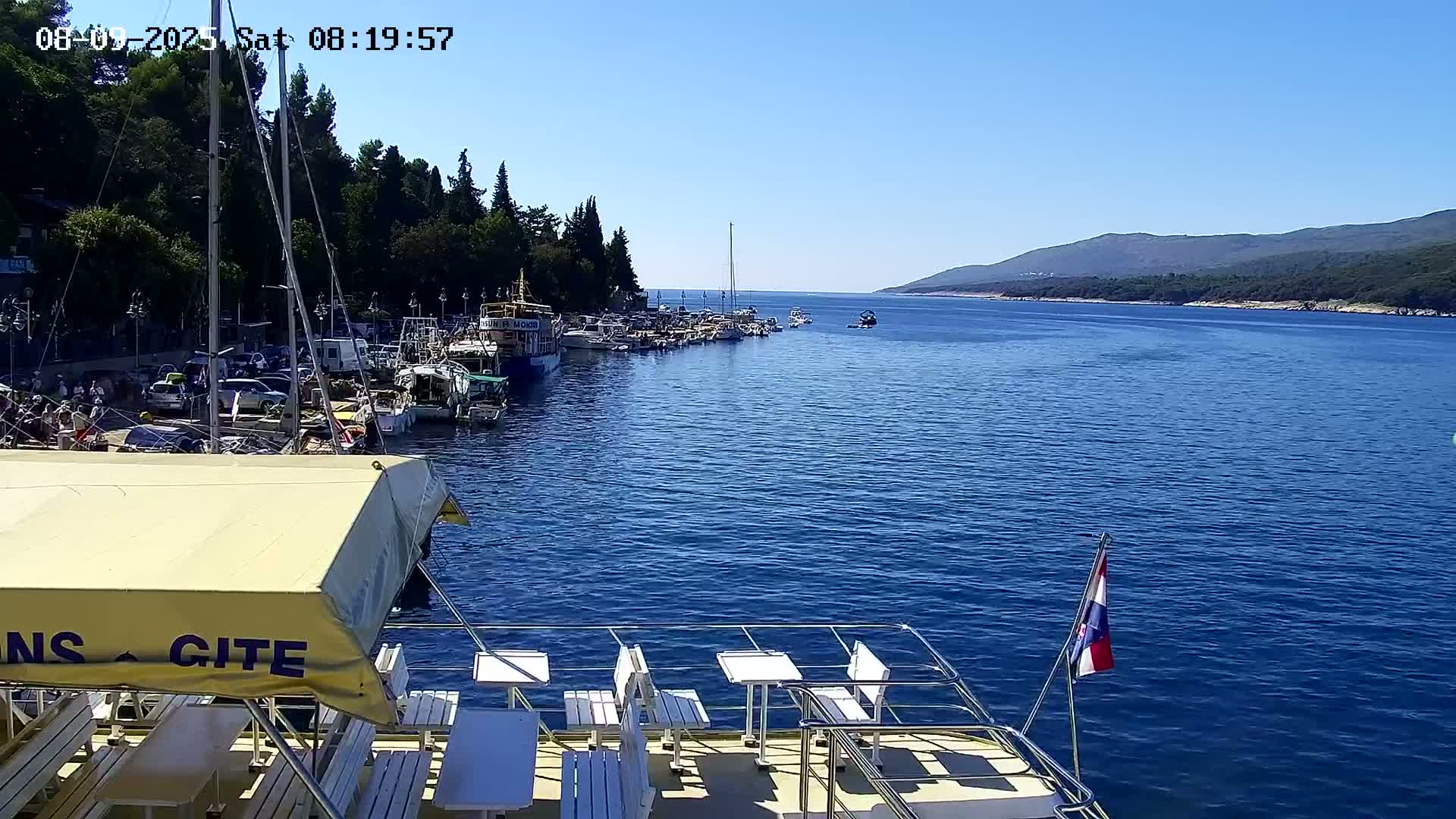 From the deck of a boat with white tables and chairs, a calm, blue sea is visible, featuring a marina filled with boats and a distant shoreline under a clear, sunny sky.