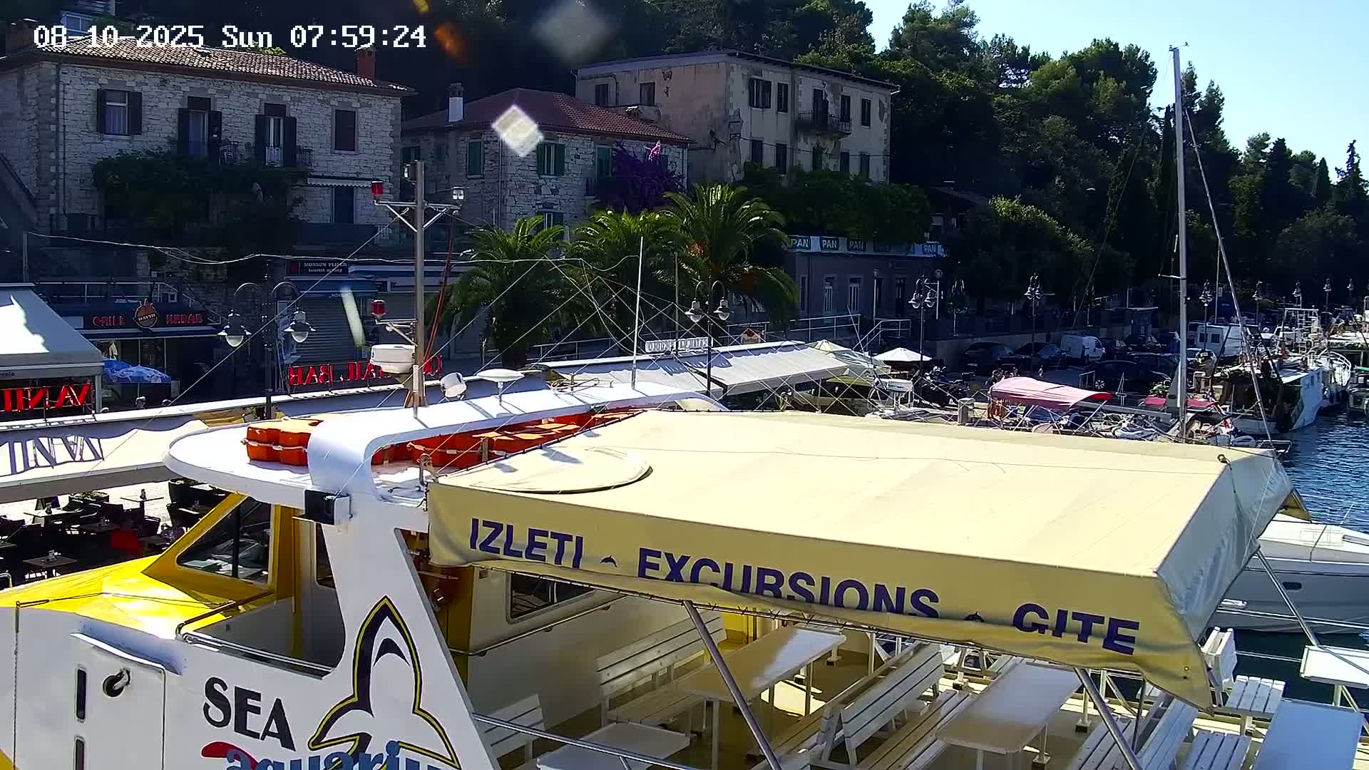 A yellow and white excursion boat with a beige awning is docked at a harbor on a sunny day, near stone buildings and other boats.