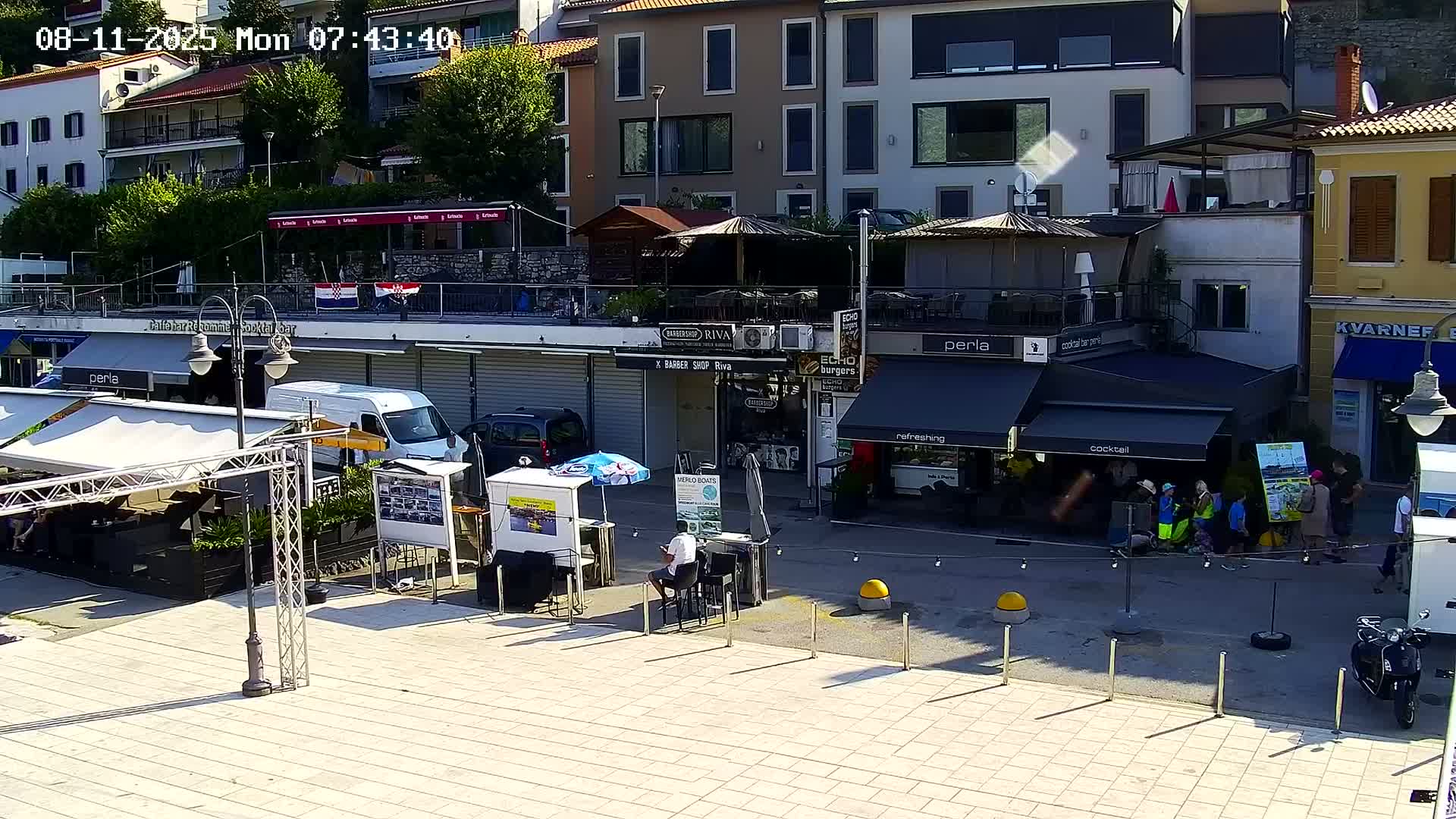 A sunny outdoor scene shows a plaza with several shops and restaurants, some with awnings, and people milling about.