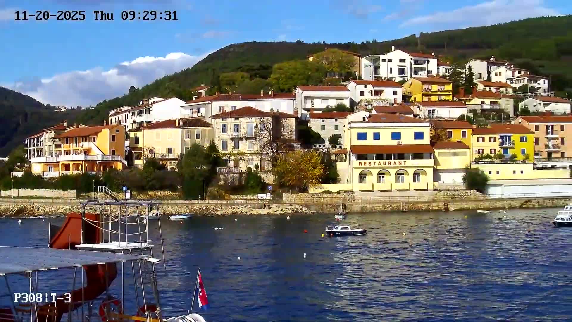 A vibrant coastal town with colorful buildings ascending a lush green hillside overlooks a calm, deep blue bay dotted with several moored boats, all under a bright, clear blue sky with scattered clouds.