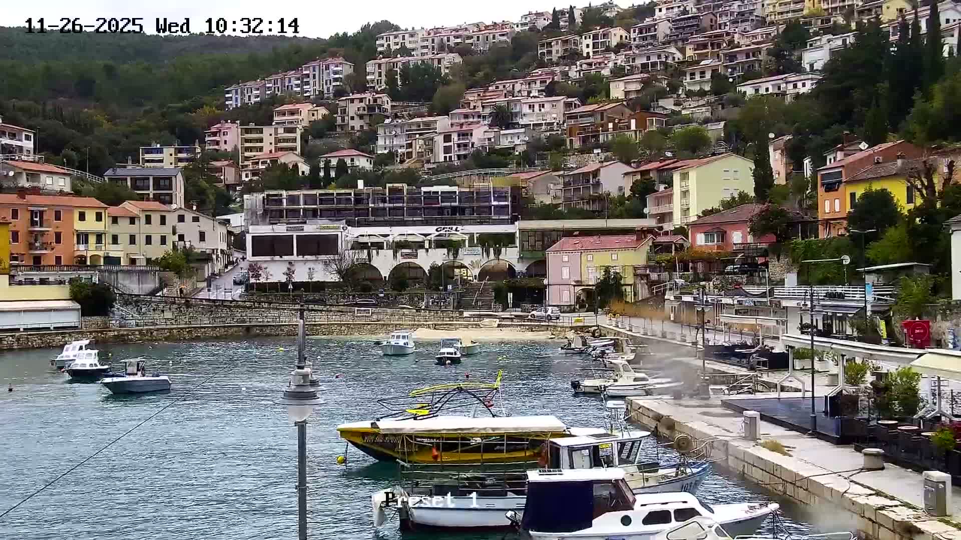 Numerous colorful houses cascade down a lush green hillside towards a calm bay dotted with boats, all observed on an overcast day.