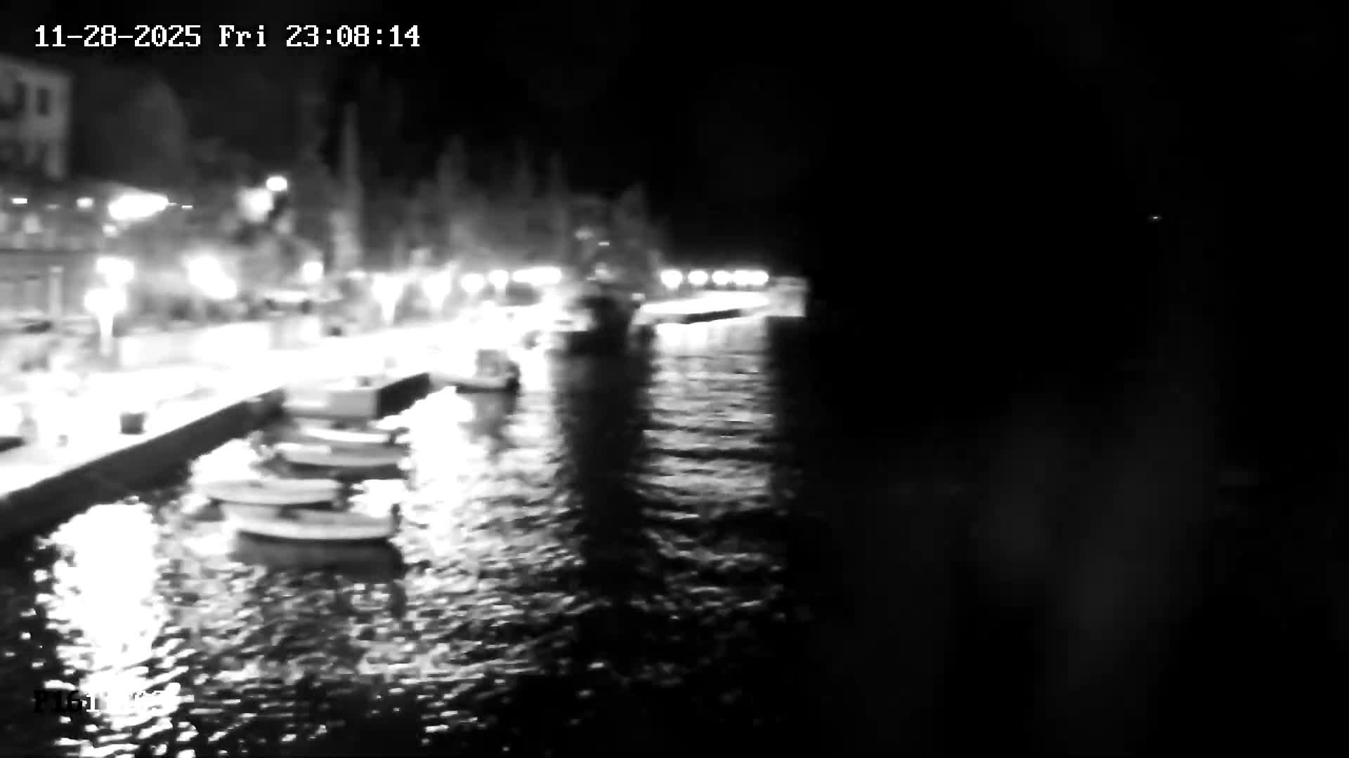 A blurry, black and white nighttime image depicts a brightly lit waterfront with several small boats moored along a quay, their reflections shimmering on the rippling water under what appears to be a clear night sky.