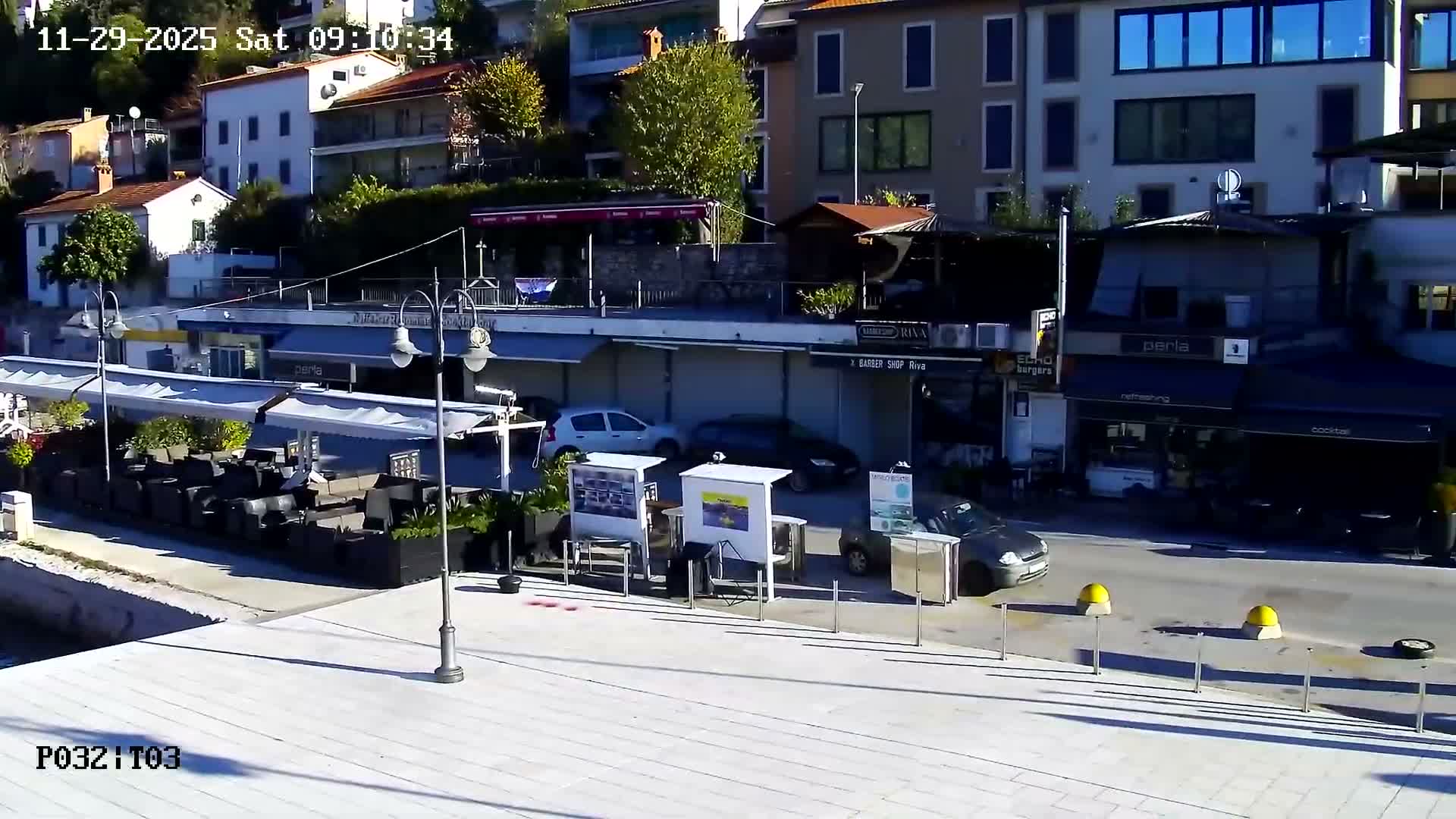 Under clear, sunny skies, a vibrant coastal scene features buildings rising on a hillside, a paved promenade with multiple outdoor cafes, and cars parked along a street.