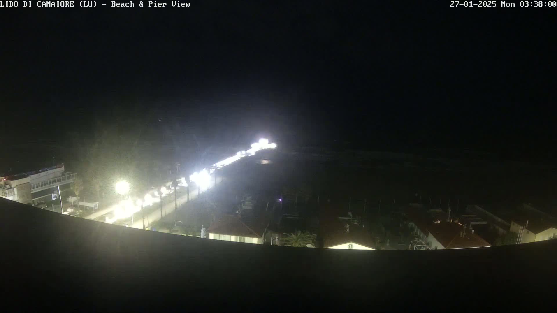 A nighttime aerial view shows a beach, pier, and a road with vehicle headlights visible, under clear skies.