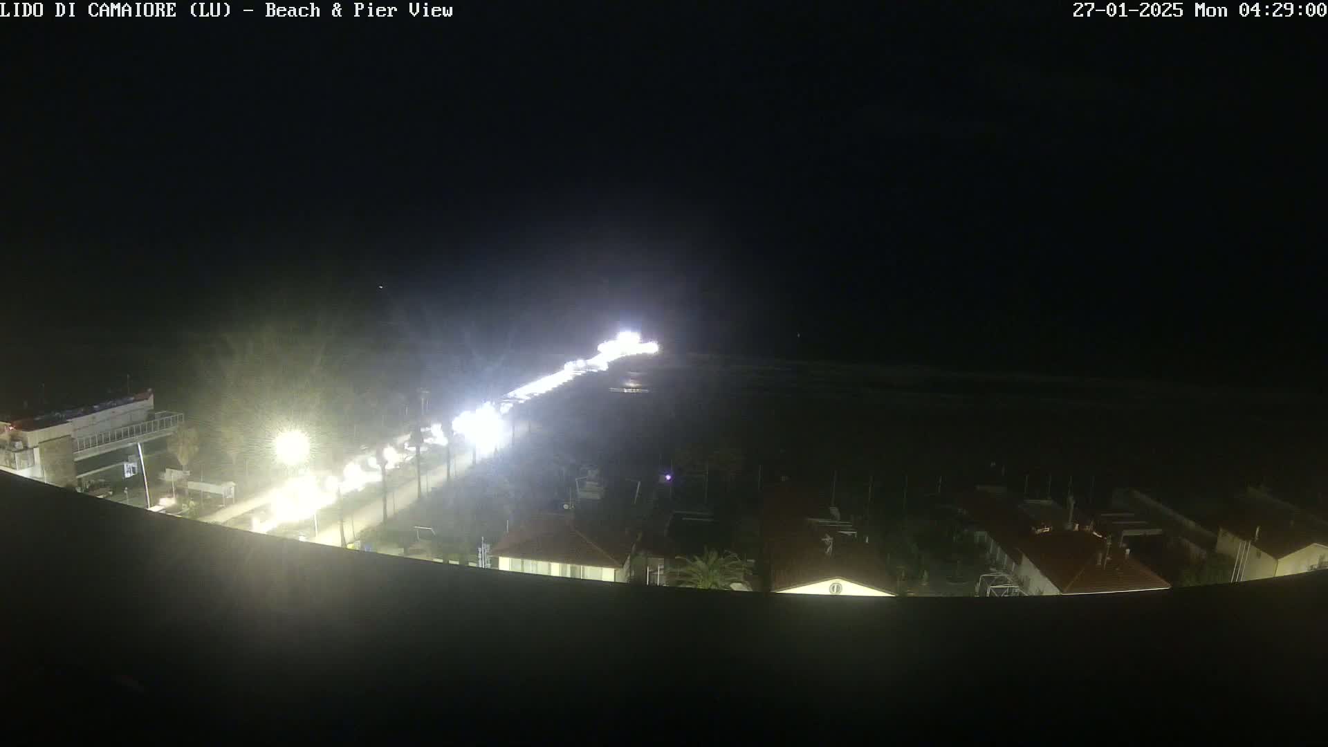 A nighttime, high-angle view shows a beach and pier with a road lined with lights and buildings visible in the distance.