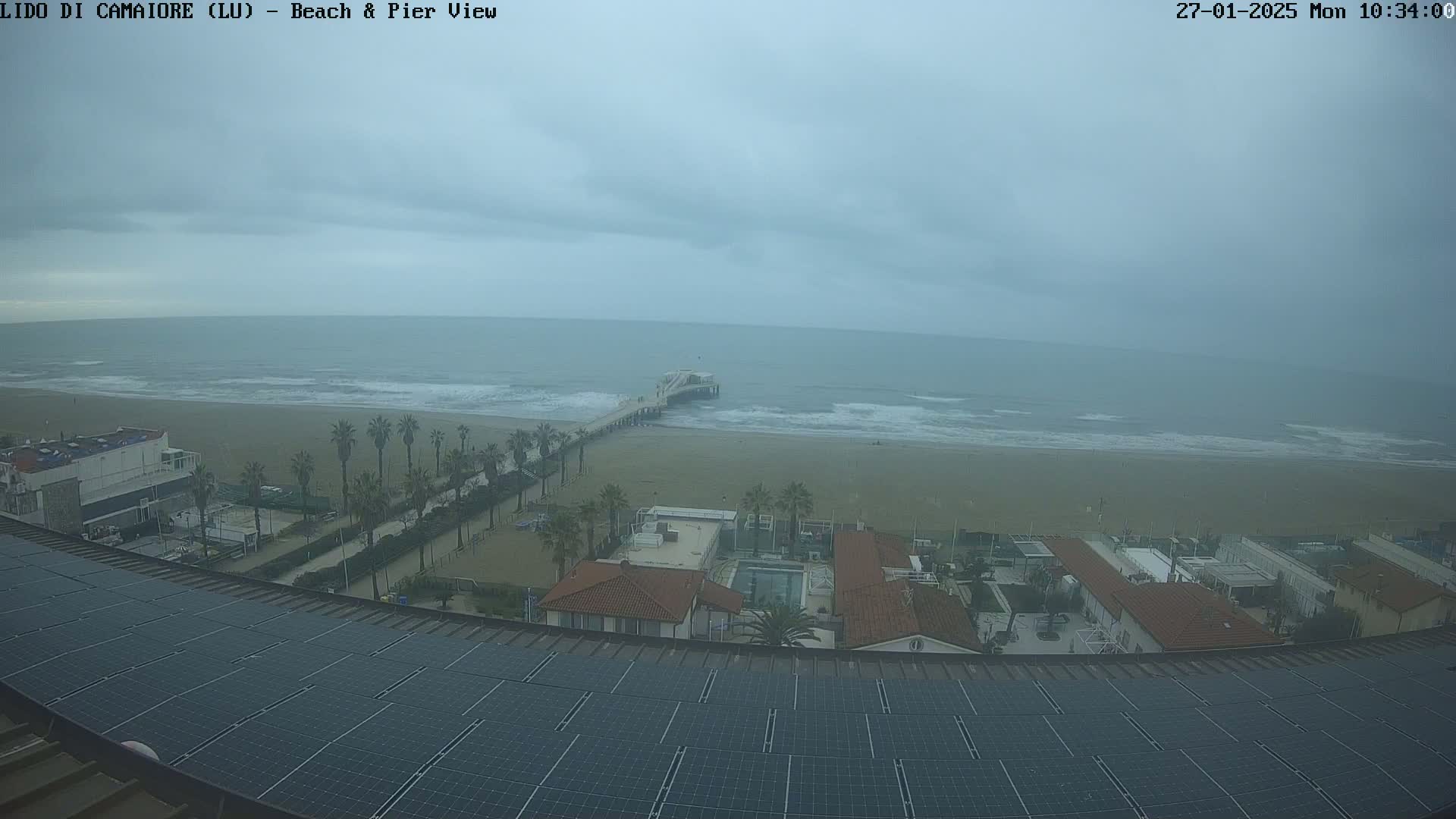 A view from a rooftop with solar panels shows an overcast day at a beach with a pier, palm trees, and buildings.