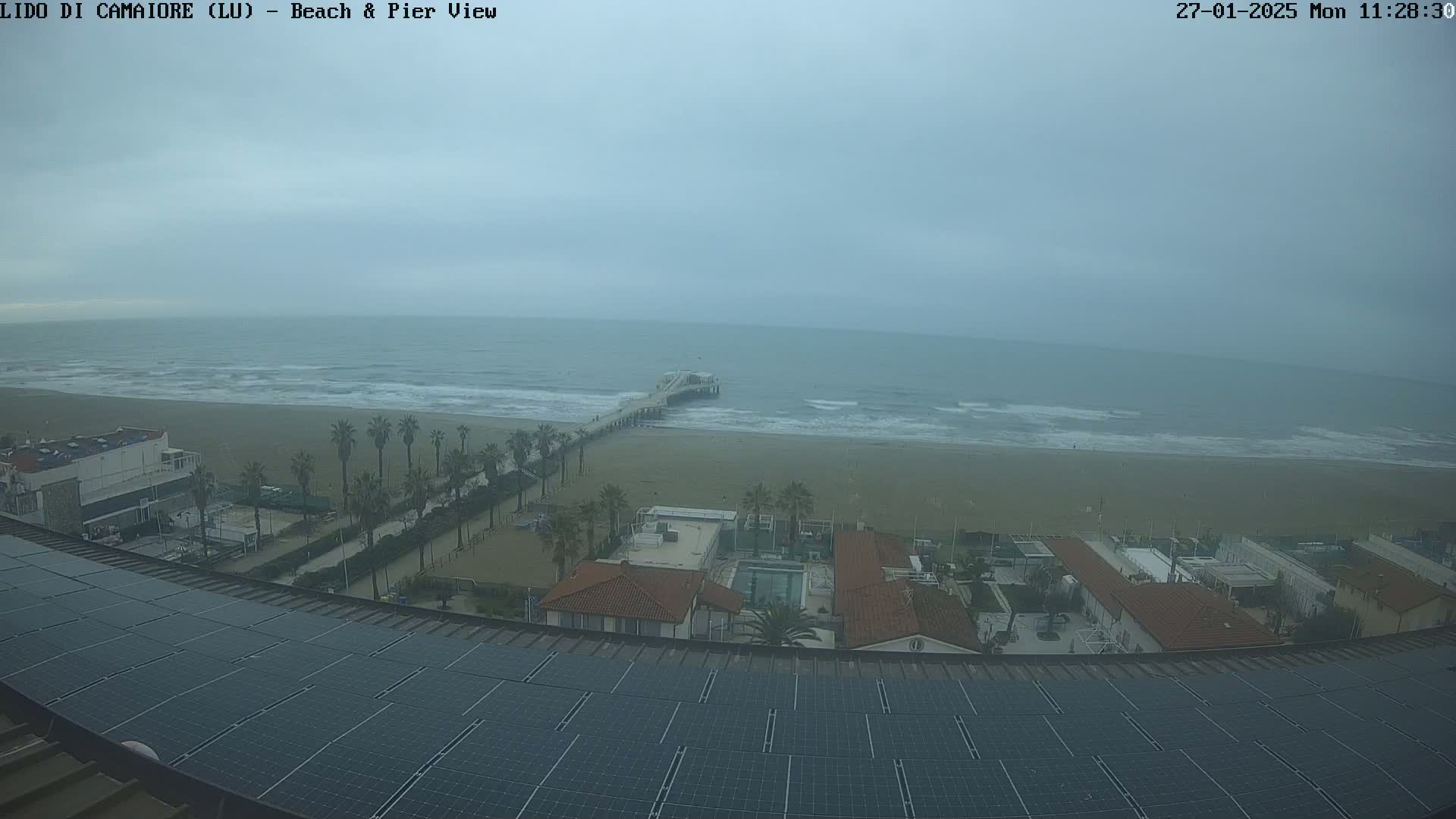 Here is a description of the image: A high-angle, long shot shows a beach with a pier under an overcast sky, viewed from a rooftop covered in solar panels.
