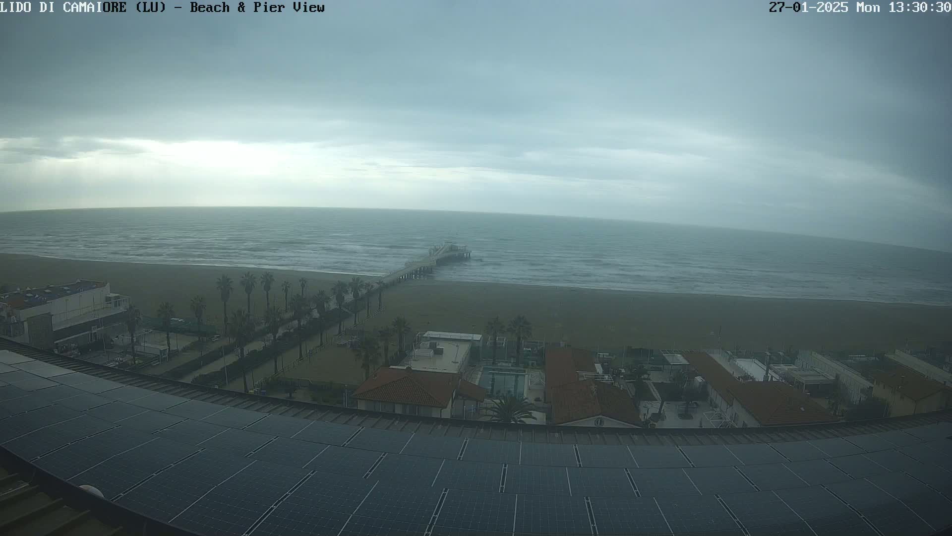 An overcast day shows a beach with a pier extending into a somewhat choppy sea, viewed from a rooftop with solar panels.