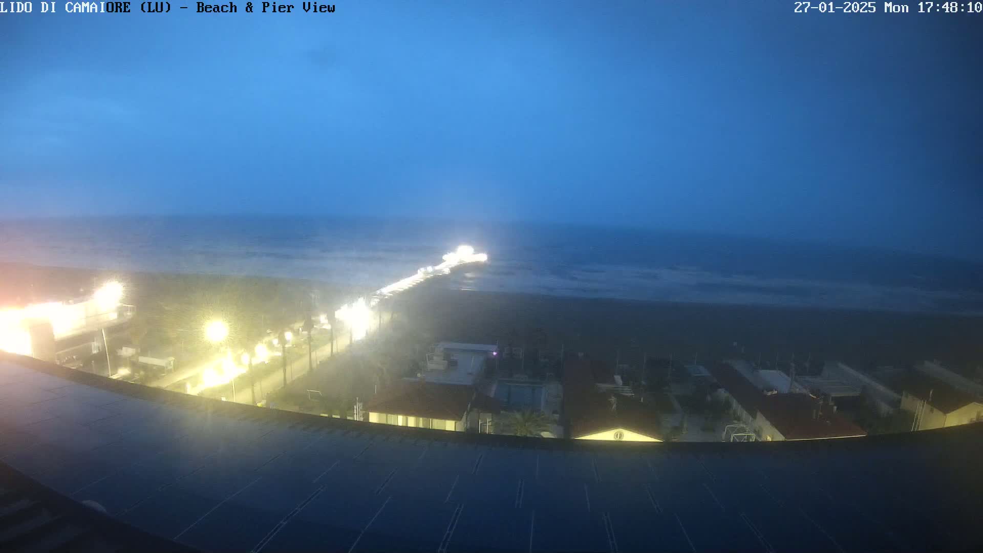 A nighttime view of a beach and pier, with a dark, possibly stormy sky and illuminated buildings and roadways.