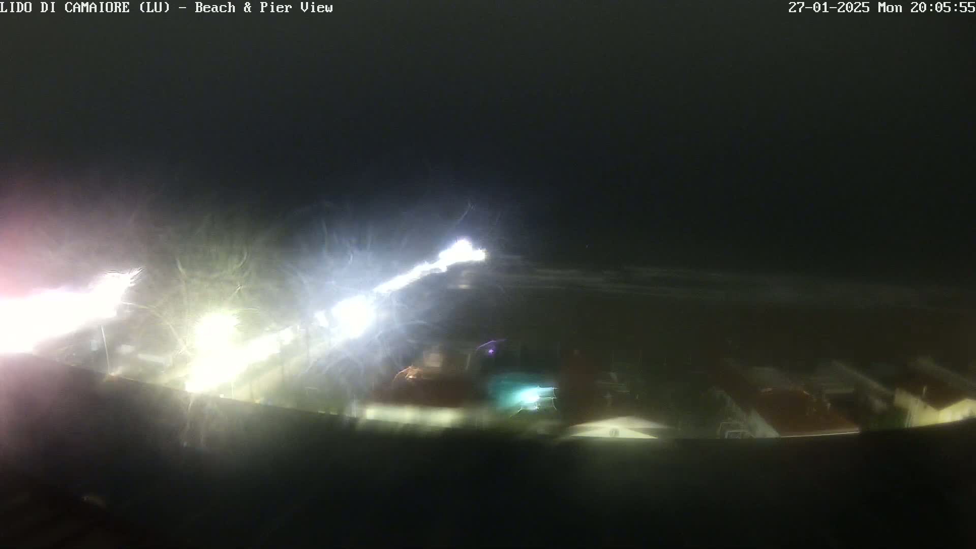 A nighttime view of a beach and pier, obscured by rain or mist, shows bright lights from vehicles or structures in the distance.