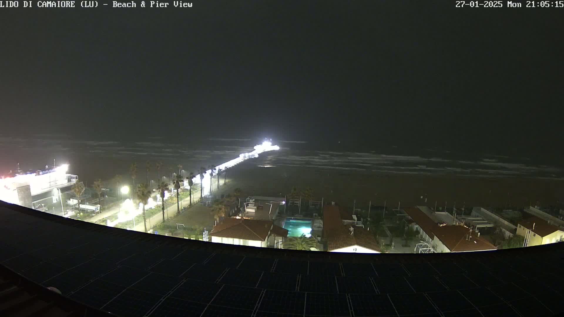 A nighttime view of a beach with a pier extending into a wavy sea under a dark, overcast sky.