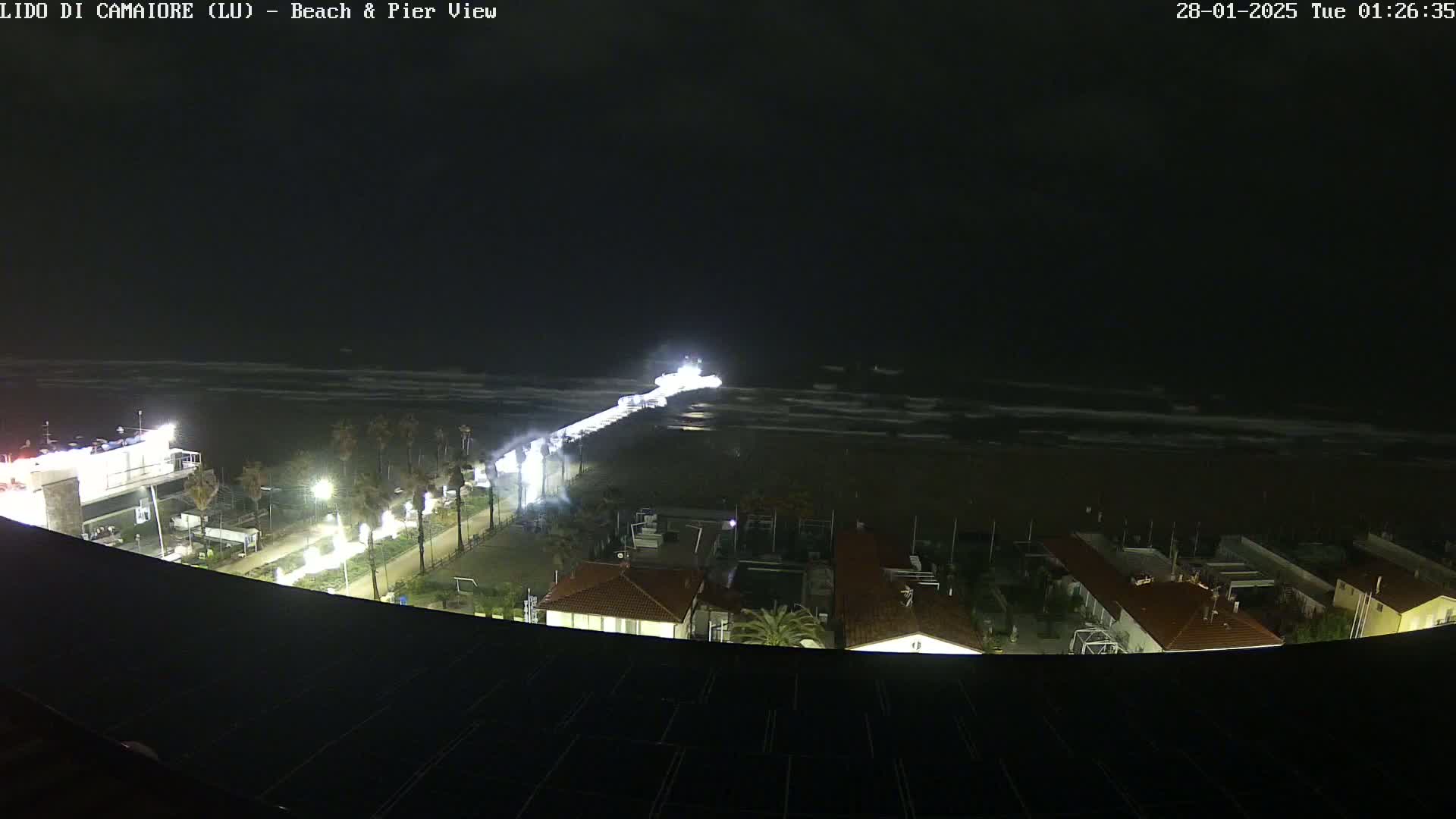 A nighttime view of a beach and pier with a brightly lit vessel at the end of the pier, and choppy waves visible in the water.