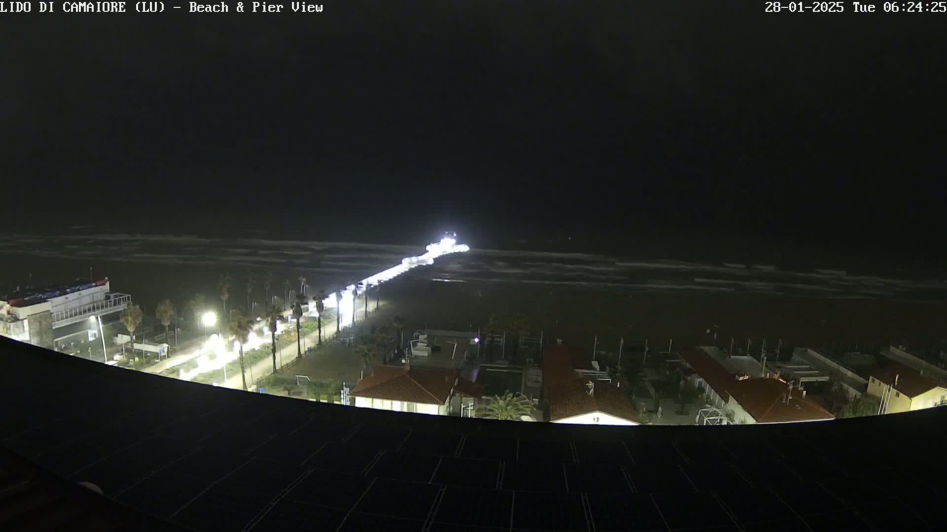 A nighttime view of a beach and pier with slightly wavy water under a dark sky, showing a coastal town with lights illuminating buildings and roads.