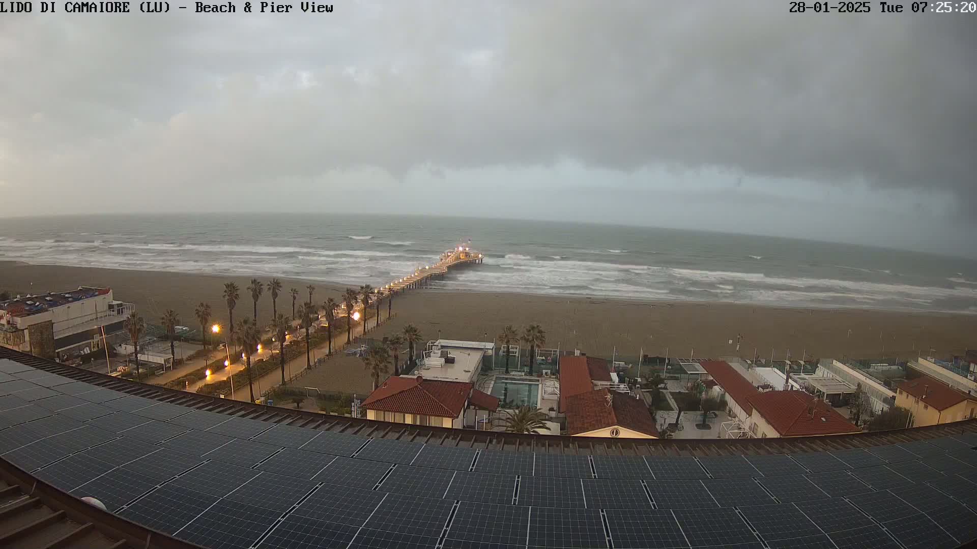 A beach with a pier and several buildings is visible from a rooftop covered in solar panels under an overcast sky, with moderate waves in the ocean.