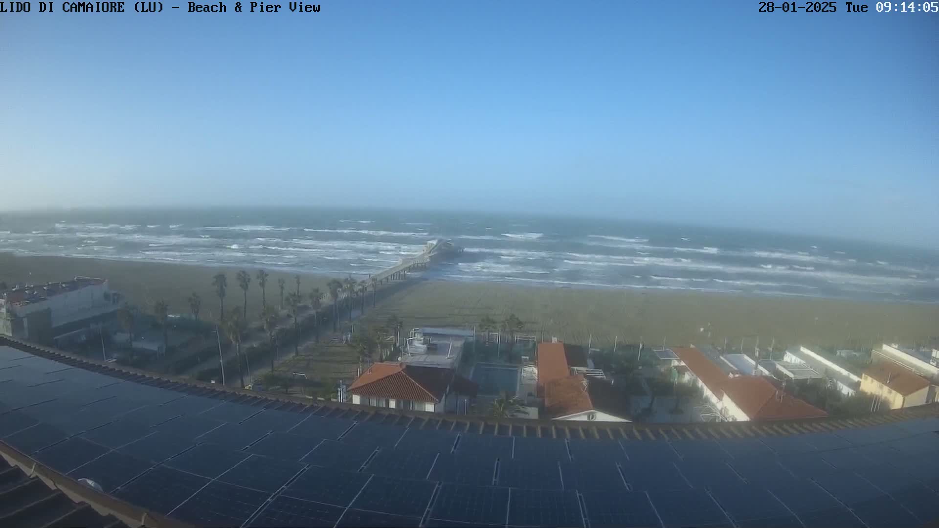 A coastal view shows a beach with choppy waves, a pier extending into the sea, buildings with red tile roofs, and a solar panel array on a rooftop under a mostly clear sky.