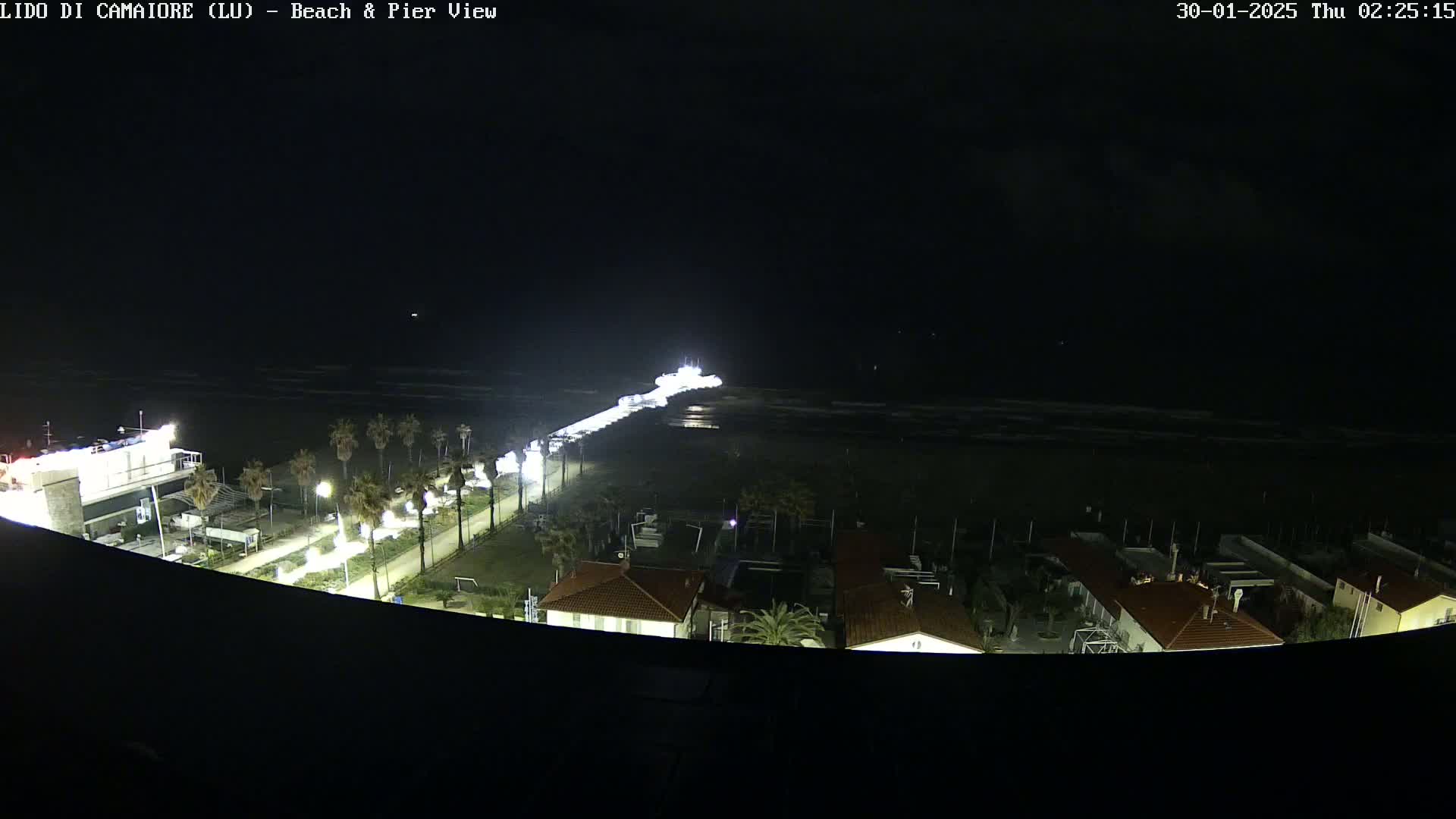 A brightly lit pier extends from a dark, nighttime beach into a calm, dark ocean, with buildings and palm trees visible on the shore.