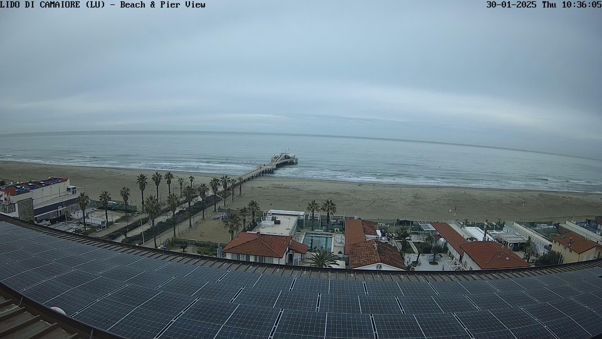 A beach with a pier extends into a calm, overcast ocean, viewed from above a rooftop covered in solar panels.