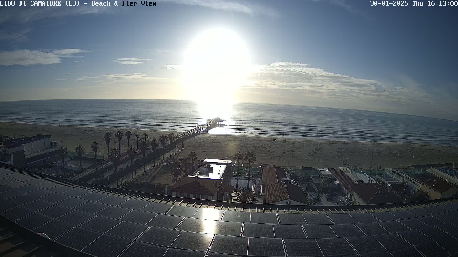 A sunny day shows a beach with a pier extending into the ocean, palm trees lining the shore, buildings and solar panels on a rooftop in the foreground.