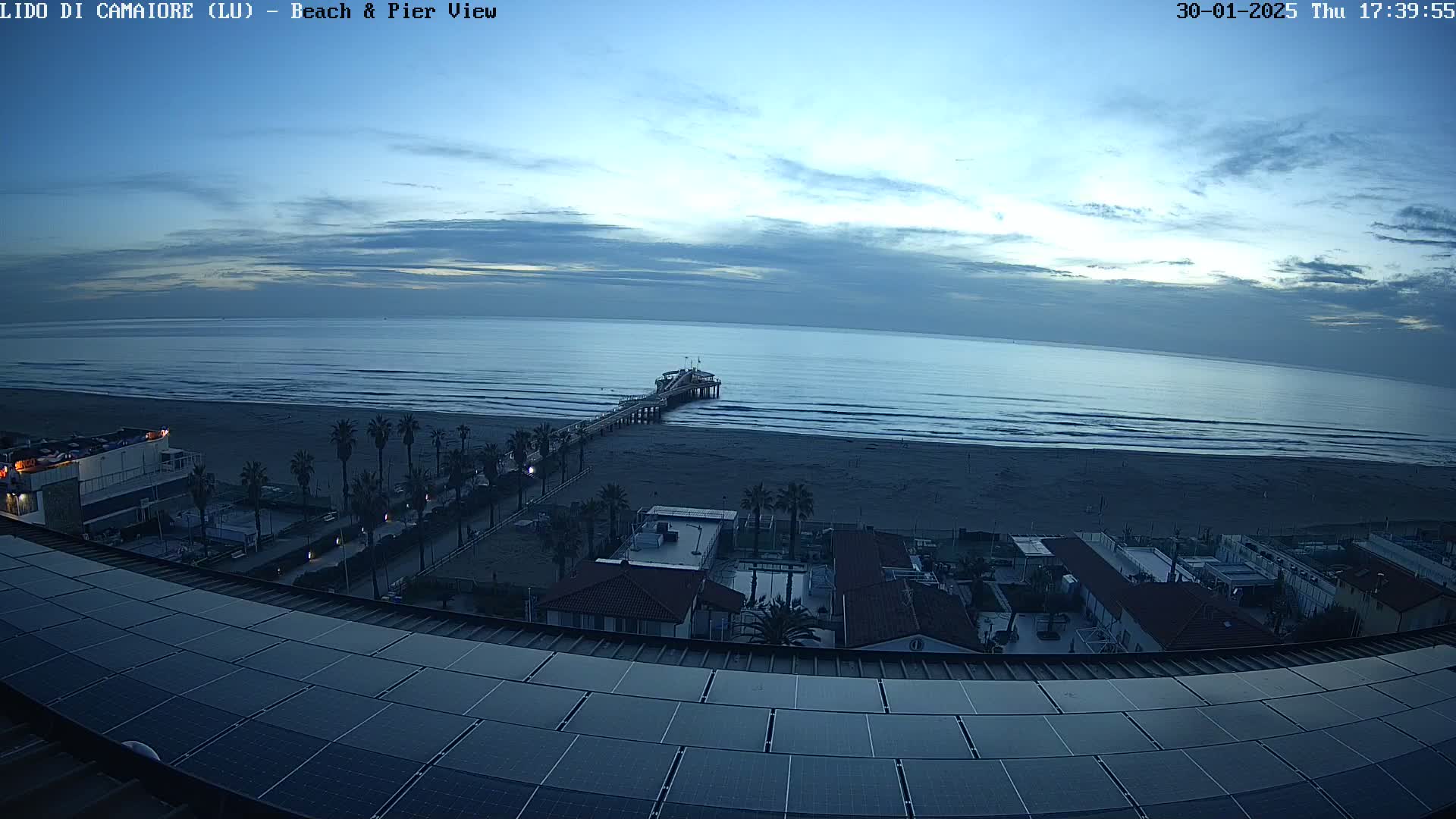 A long pier extends into a calm, blue ocean under a twilight sky, viewed from a rooftop with solar panels overlooking a beach and beachfront buildings.