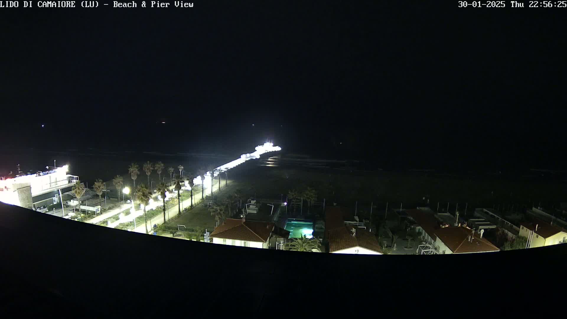 A nighttime aerial view shows a pier extending from a brightly lit beachfront area into a dark ocean, with palm trees lining the road leading to the pier and buildings visible behind it.
