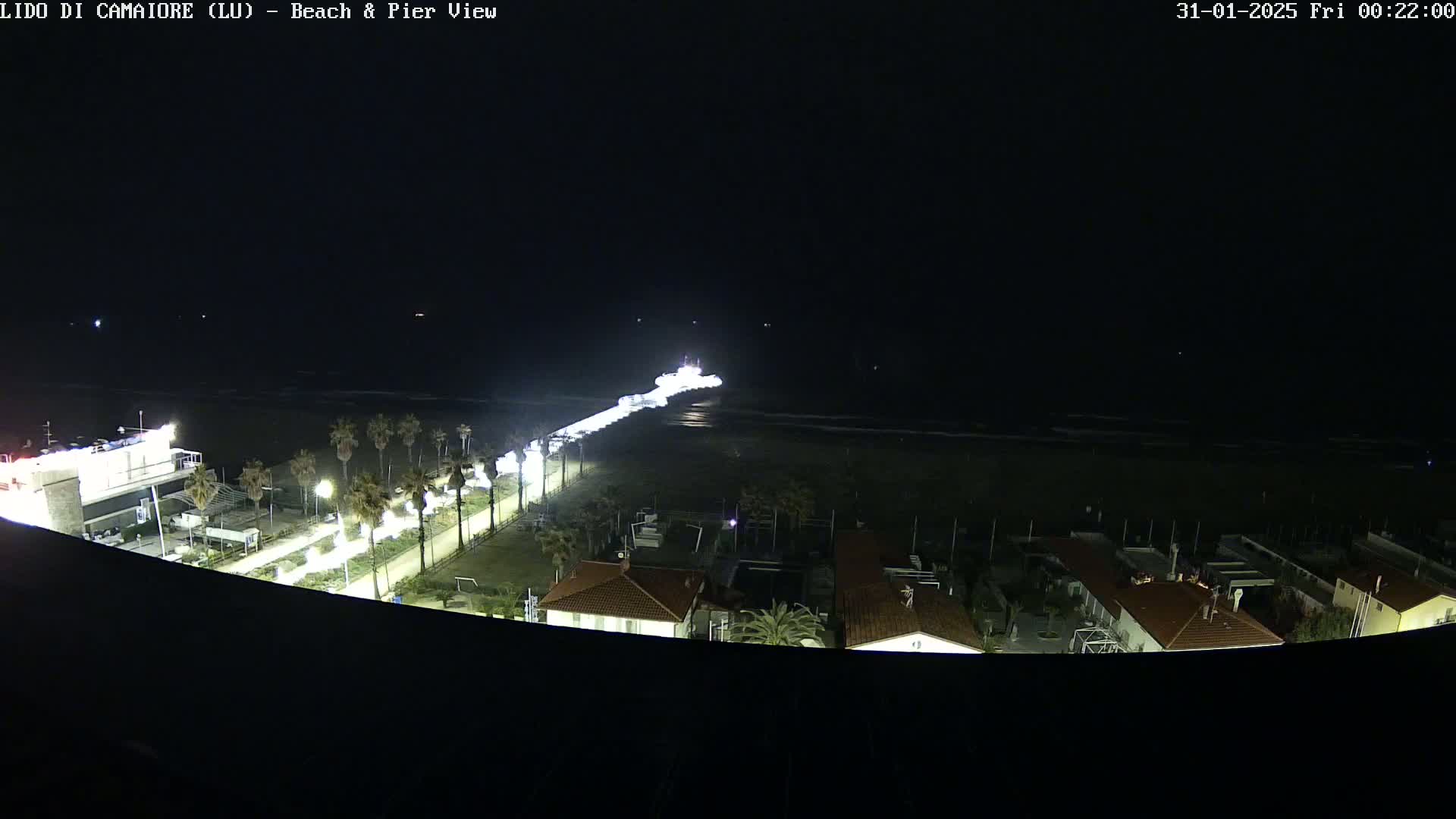 A nighttime aerial view shows a pier extending into a dark ocean, illuminated by lights, with palm trees and buildings visible along the shoreline.
