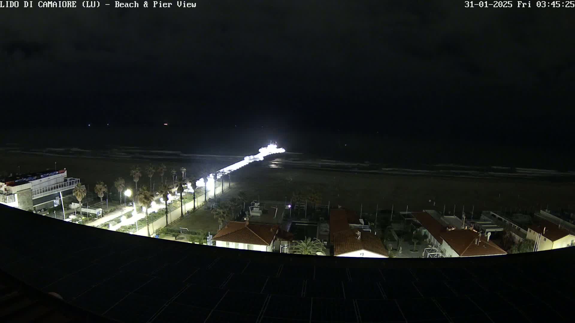 A nighttime view of a beach and pier, with a brightly lit area on the pier and calm ocean waves.
