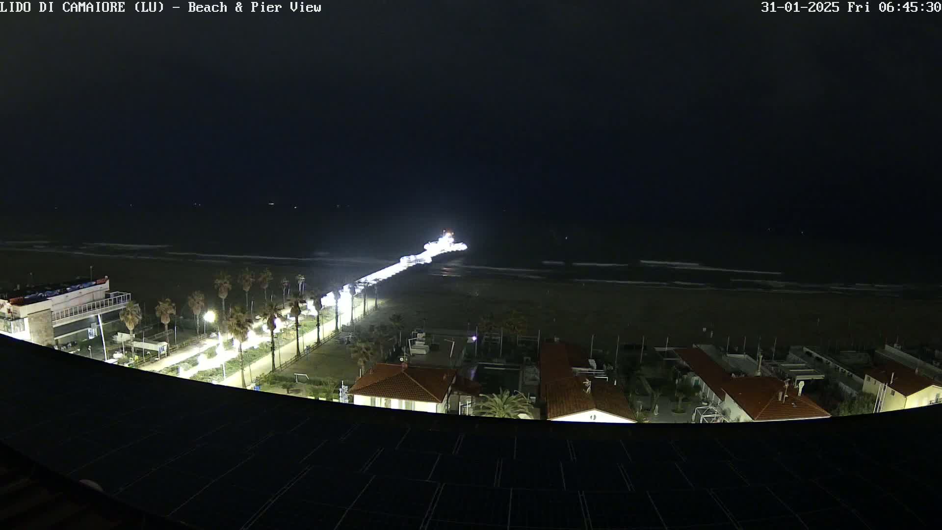 A nighttime view of a beach and pier with a dark ocean and illuminated beachfront buildings.