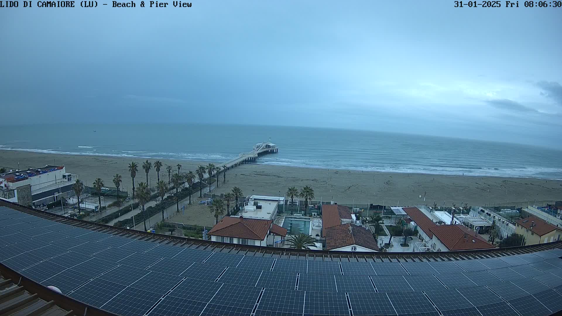 A long sandy beach with a pier extends into a calm, overcast ocean, viewed from a rooftop covered in solar panels.