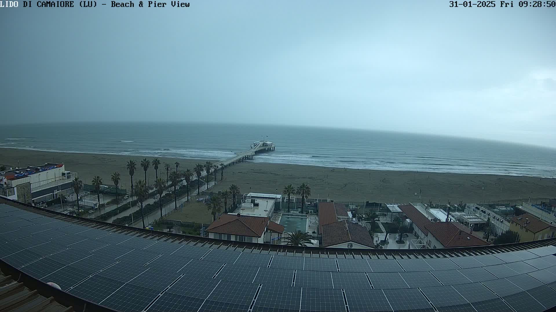 A beach with a pier and several buildings is viewed from above on an overcast day, showing a row of solar panels on a roof in the foreground.