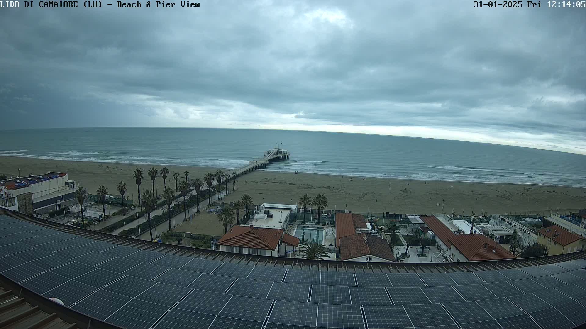 A beach with a pier is visible under an overcast sky from a rooftop covered in solar panels.