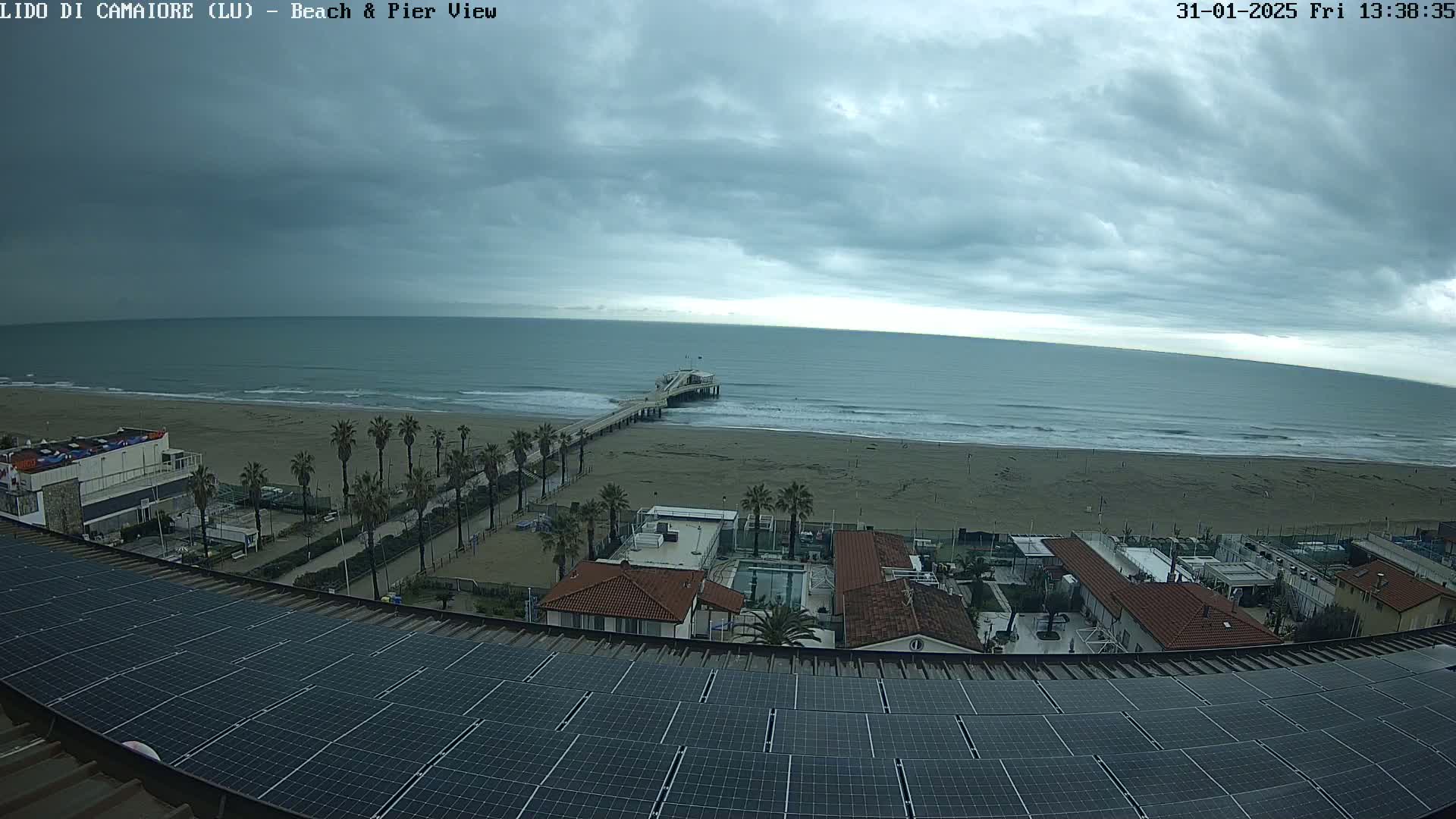 A beach with a pier extends into a wavy ocean under a cloudy sky, viewed from above a rooftop covered in solar panels.