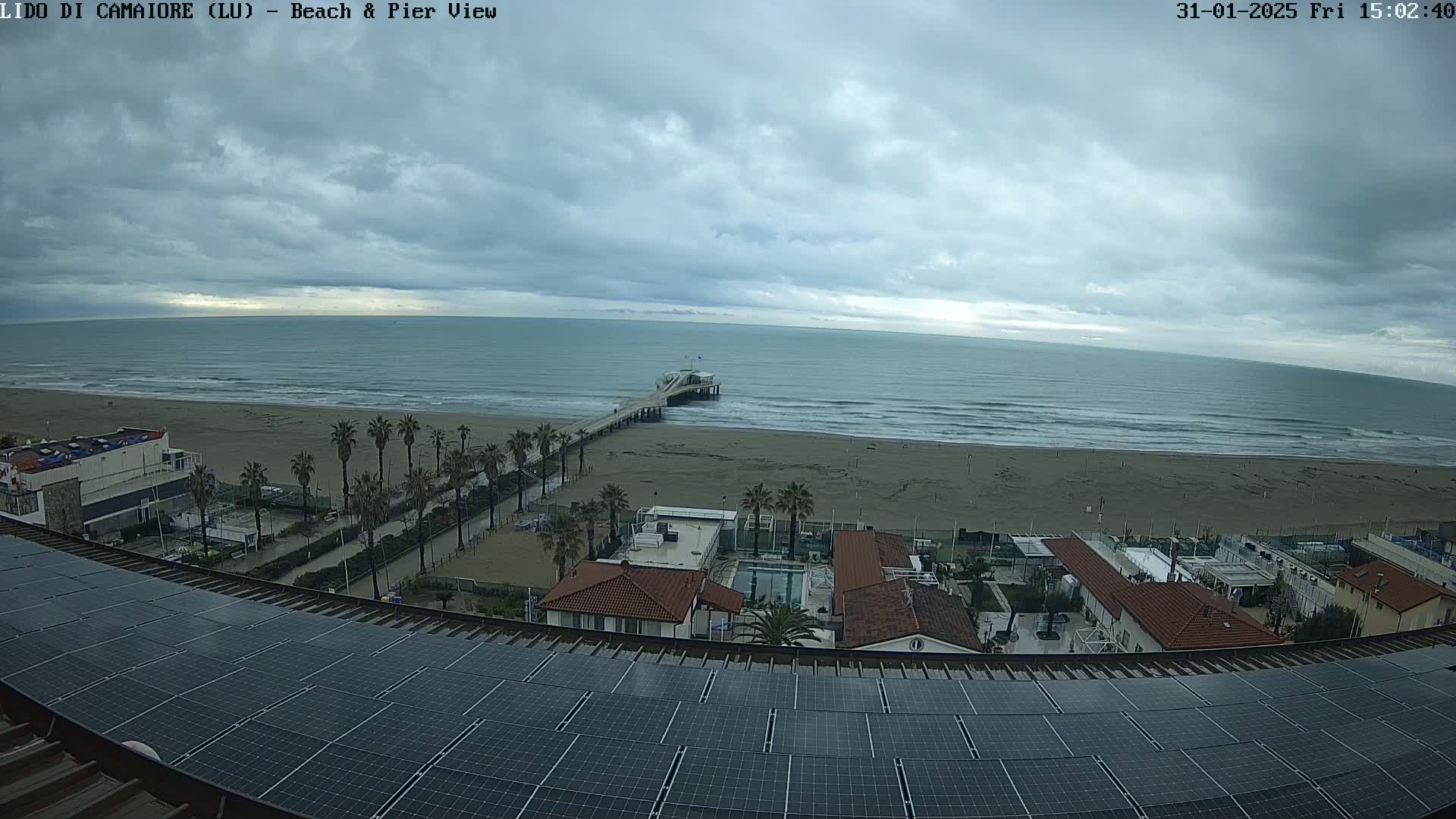 A beach with a pier is viewed from a rooftop covered in solar panels under an overcast sky.