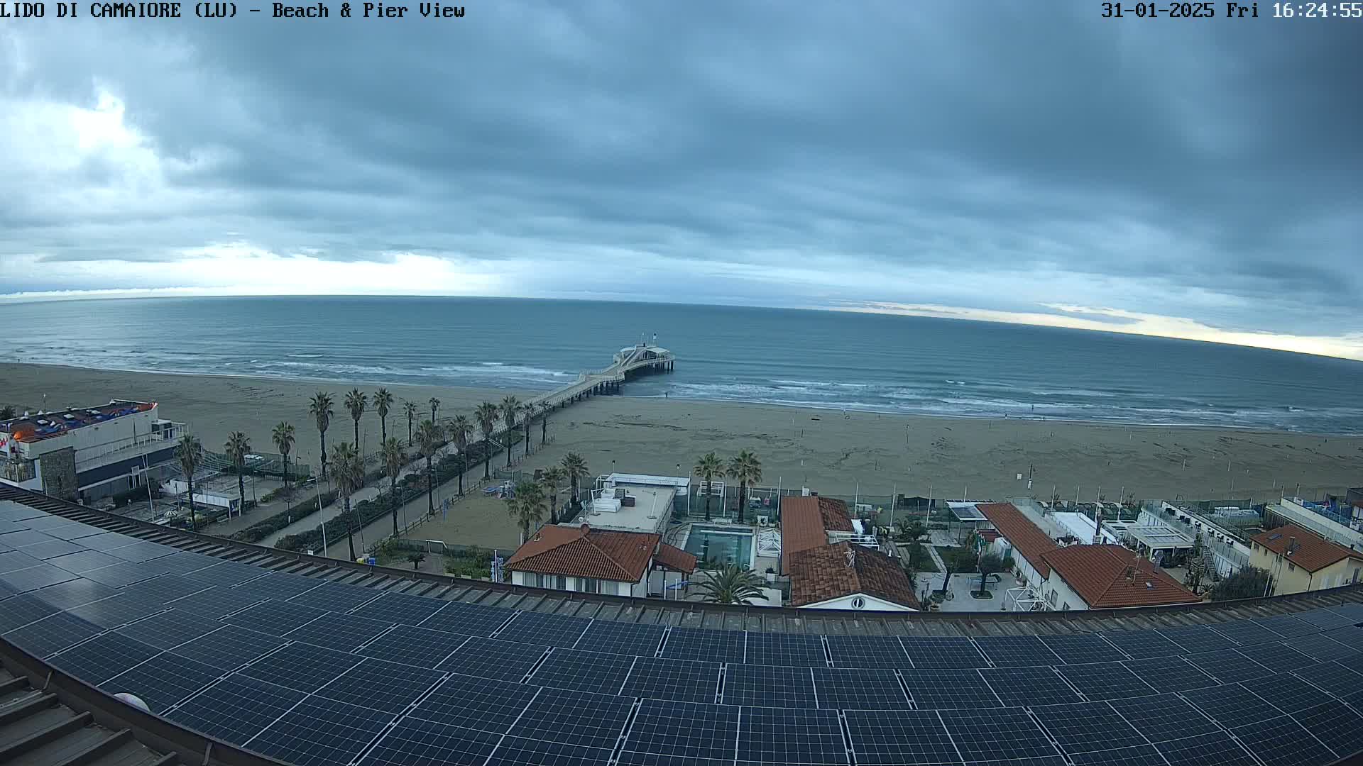 A beach with a pier extends into a calm sea under a cloudy sky, viewed from above a rooftop covered in solar panels.
