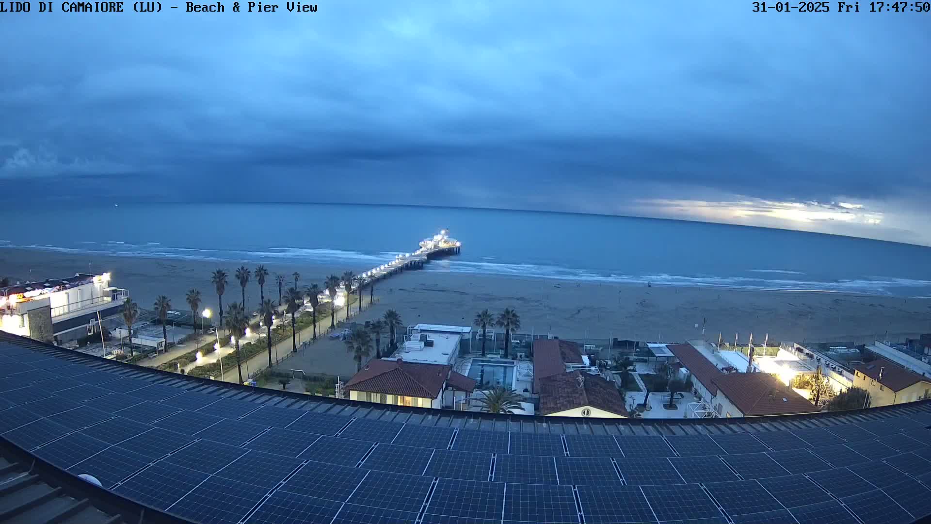 A view from a rooftop with solar panels shows a beach, pier, and ocean under an overcast sky at dusk.