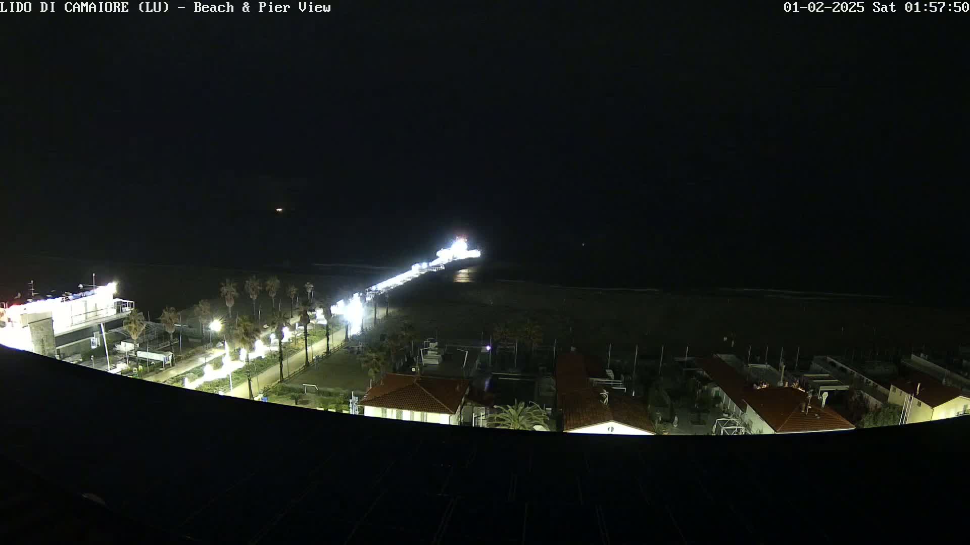 A nighttime aerial view shows a brightly lit pier extending from a shoreline into a dark ocean, with buildings and palm trees visible along the coast.