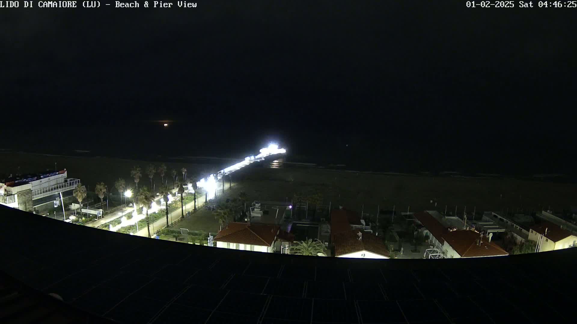 A nighttime aerial view shows a brightly lit pier extending into a dark ocean, with a beach and buildings visible below under a clear, dark sky.