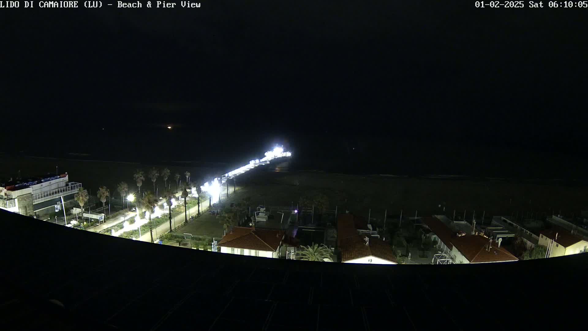 A brightly lit pier extends from a dark, nighttime beach into the ocean, with buildings and palm trees visible along the shoreline.